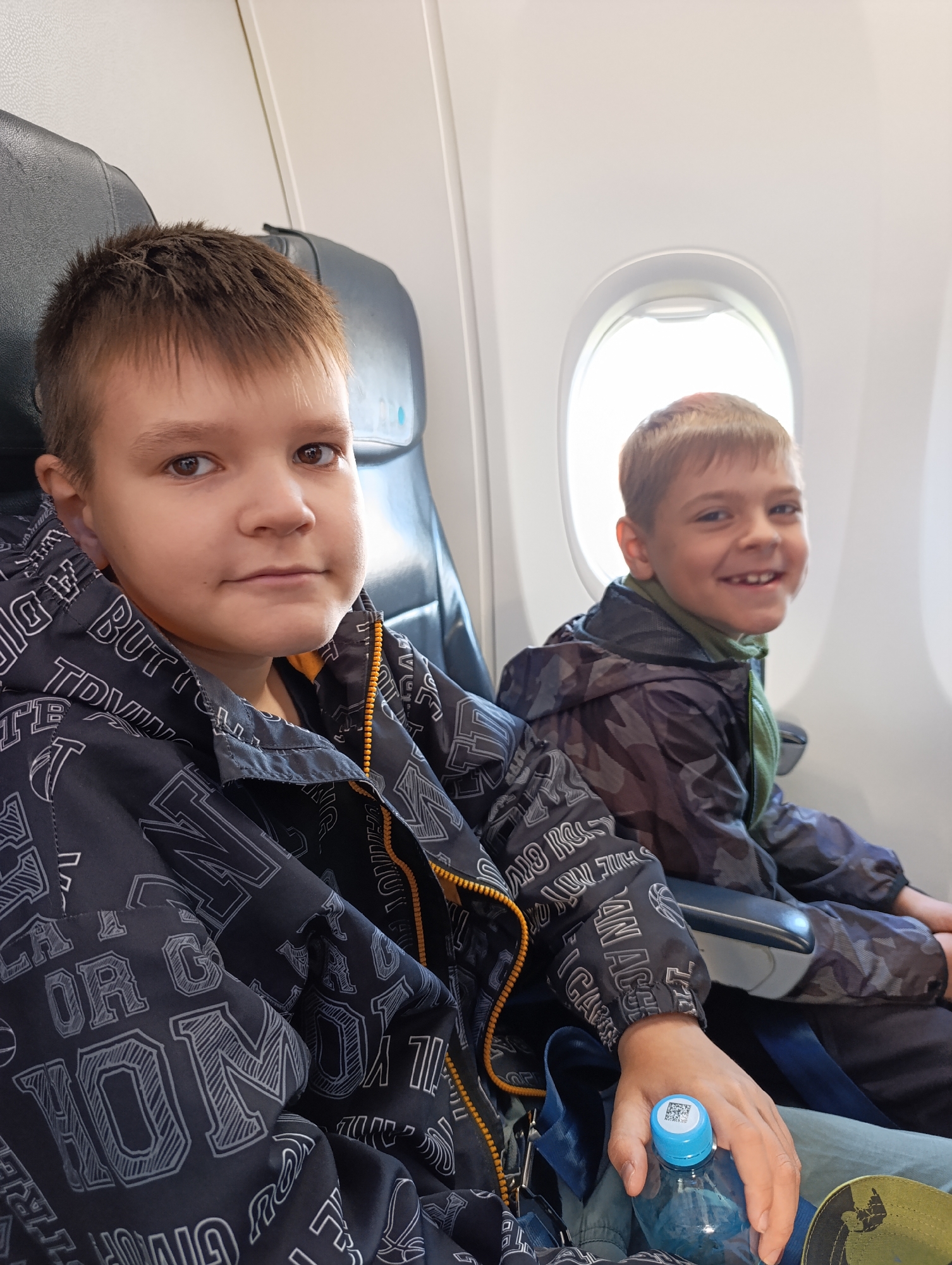 Two young boys are seated inside an airplane cabin, captured in a bright, daylight setting. The boy in the foreground is positioned closer to the camera, looking straight ahead with a neutral, calm expression. He has short brown hair and dark eyes, and is wearing a dark jacket adorned with numerous light-colored letter patterns and an orange zipper. Visible text fragments on his jacket include "BUT", "TE", "SHINE", "SIDI GIV", "BIGARR", "AN ACCE", "OP", "OL", "NOL", and "OR C". His right hand is holding a blue plastic water bottle with a blue cap that has a QR code sticker.
Behind him and to his right, closer to the airplane window, sits a second boy. This boy is smiling broadly and happily, showing his teeth, and has lighter brown hair. He is wearing a dark, possibly camouflage-patterned jacket over a green top. Bright natural light floods through the distinctive oval airplane window next to him. The seats in the cabin are dark-colored. The image captures a moment during air travel, likely departing from or arriving in Bolshoye Savino, Russia, given the context.