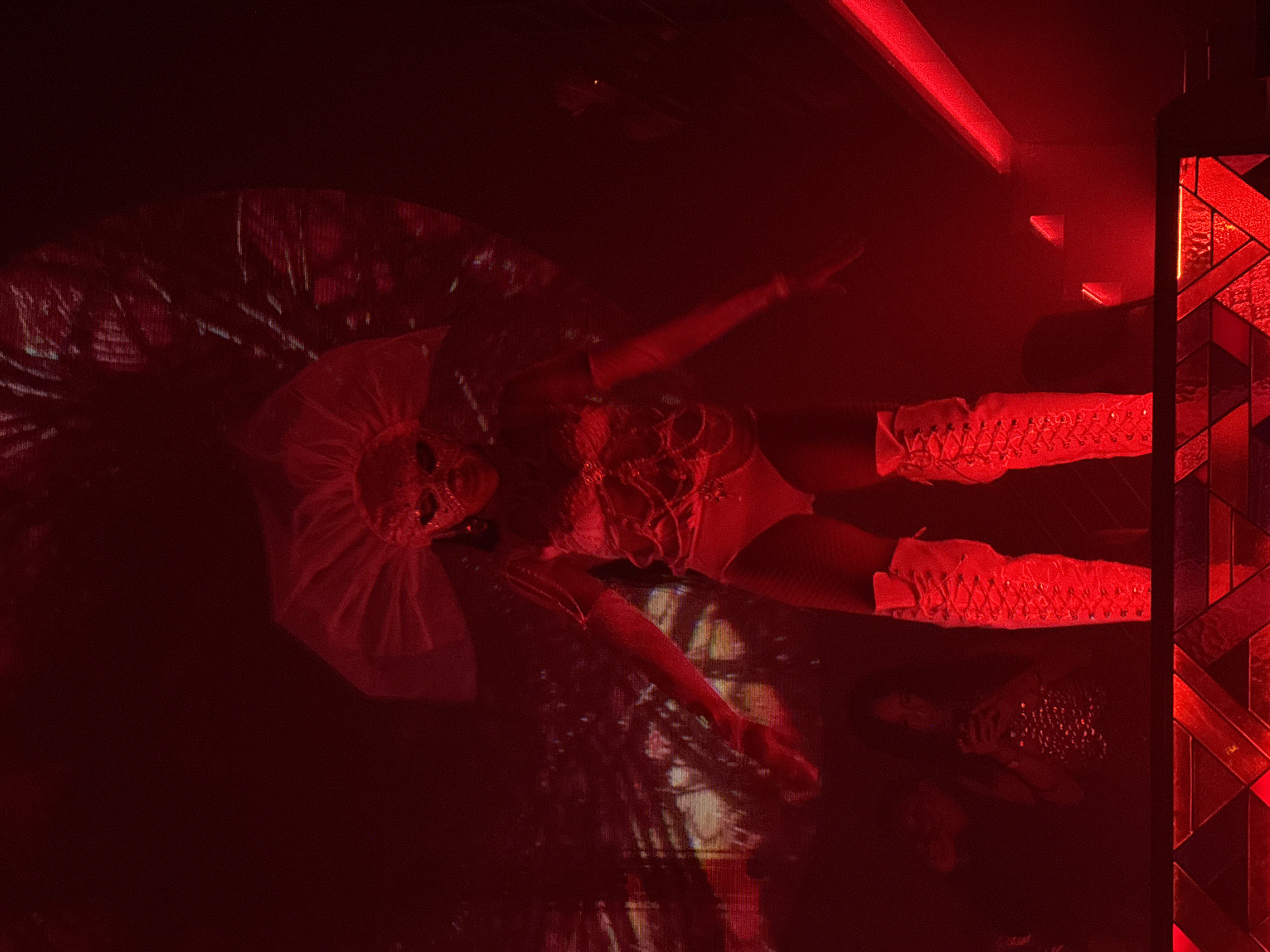 A performer, dressed in a glittery, red, sequined outfit, a mask, and thigh-high white lace-up boots, stands on a raised platform in a dimly lit room, illuminated by red lighting. The performer wears a large, fan-like headdress. Behind the performer, a large screen displays a blurry, abstract pattern. In the background, other individuals are visible in the shadows. The environment appears to be a nightclub or performance venue in North Kuta, Indonesia.