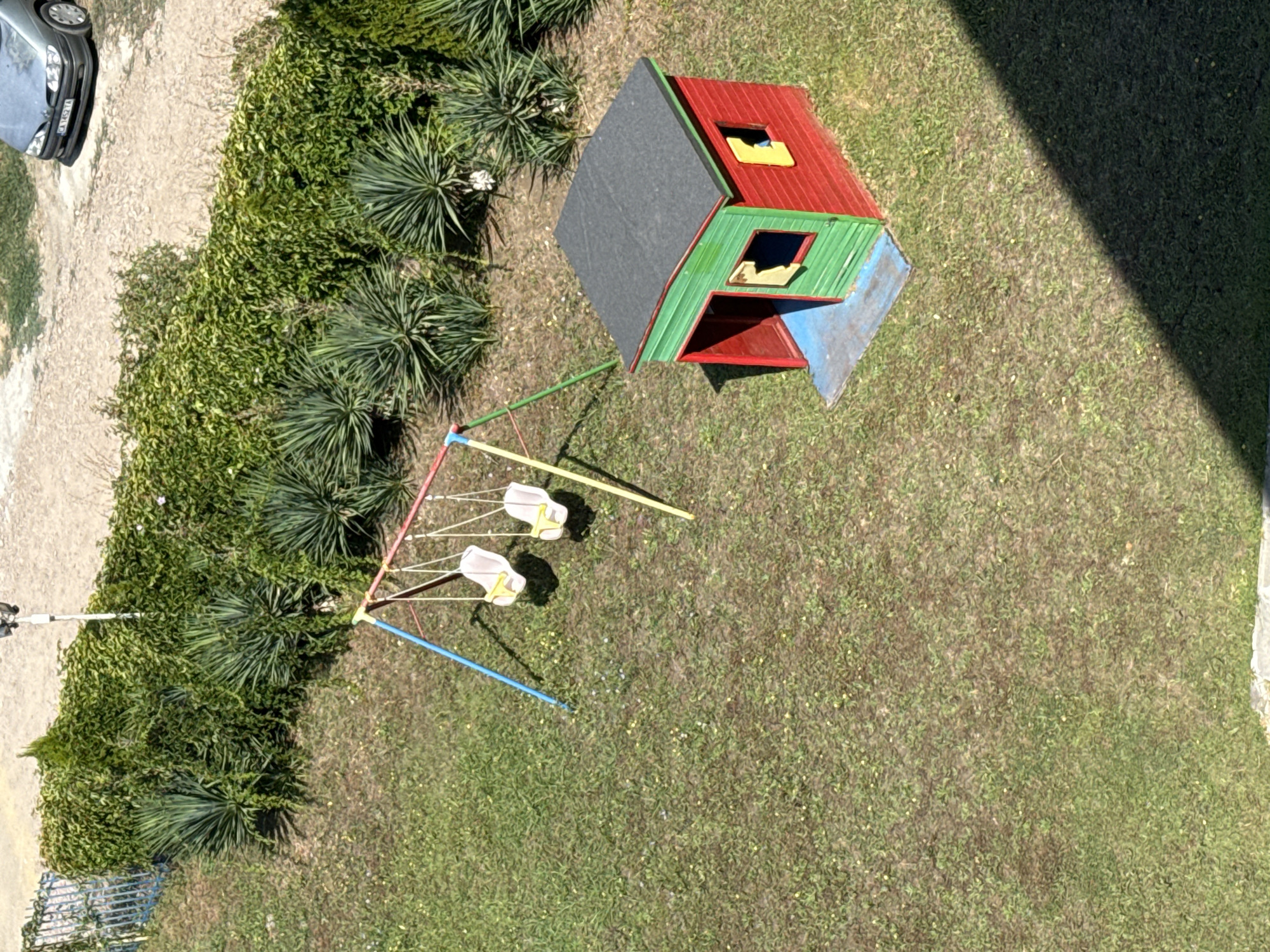 An aerial view of a small playground in Pomorie, Bulgaria shows a red and green playhouse with yellow window frames and a blue base, situated on a grassy area. Adjacent to the playhouse is a swing set with two white swings hanging from a multi-colored frame. The playground is bordered by a hedge and some yucca plants. In the background, a gravel path leads to a parked silver car. The scene is illuminated by sunlight, casting shadows on the grass.