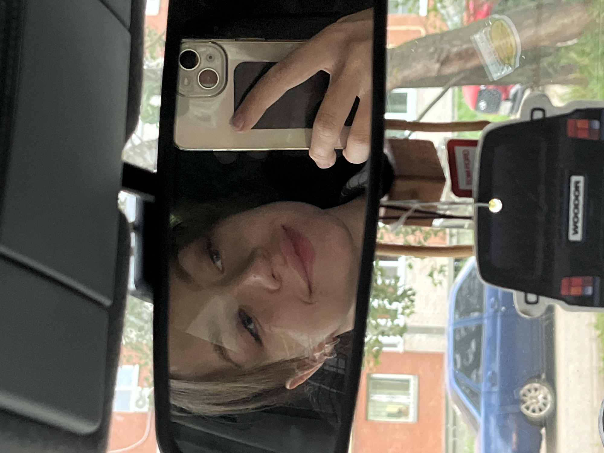 The media file is a selfie taken in a car's rearview mirror. The subject is a young person, likely female, with blonde hair, looking directly at the camera. Her face is reflected, showing her eyes, nose, and lips, which appear to be lightly colored with lipstick. She is holding a smartphone in her right hand, with her fingers curved around the back of the device. The phone is silver and has a visible camera array.
The setting is inside a car, as indicated by the rearview mirror and the dark interior visible in the foreground. Outside the car, through the windshield and side windows, a blurred scene of a residential area can be seen. This includes buildings with windows, trees with green foliage, and a blue car parked in the background. The lighting suggests it is daytime, likely morning or afternoon, with overcast or diffused light. The overall mood is casual and personal, typical of a selfie.
There is some visible text in the image. On the side of the car in the background, a black object has the word "WOOOD" in white letters. Another visible piece of text, partially obscured, appears to be in a red rectangular frame and might be a warning or informational sign, possibly reading "STOP ROAD" or similar. The location context is Chaikovsky, Russia, which is consistent with the architecture and vehicle types that might be found there.