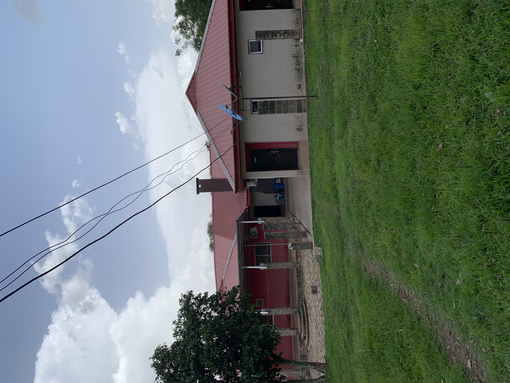 The image depicts a building with a red, gabled roof, situated in a grassy area. The building has a red-colored section on the left with a covered walkway supported by brick pillars. This section has windows and a set of steps leading to a paved area. To the right, the building is painted a light beige color and features several windows and doors. Two satellite dishes are mounted on the roof. Overhead power lines crisscross the sky, which is partly cloudy. A tree with dark green foliage stands to the left of the building. The ground in front of the building is covered in green grass, with a dirt path winding through it. The scene appears to be located in Jos, Nigeria, though no specific landmarks are visible to confirm this. There are no people or animals present in the image.