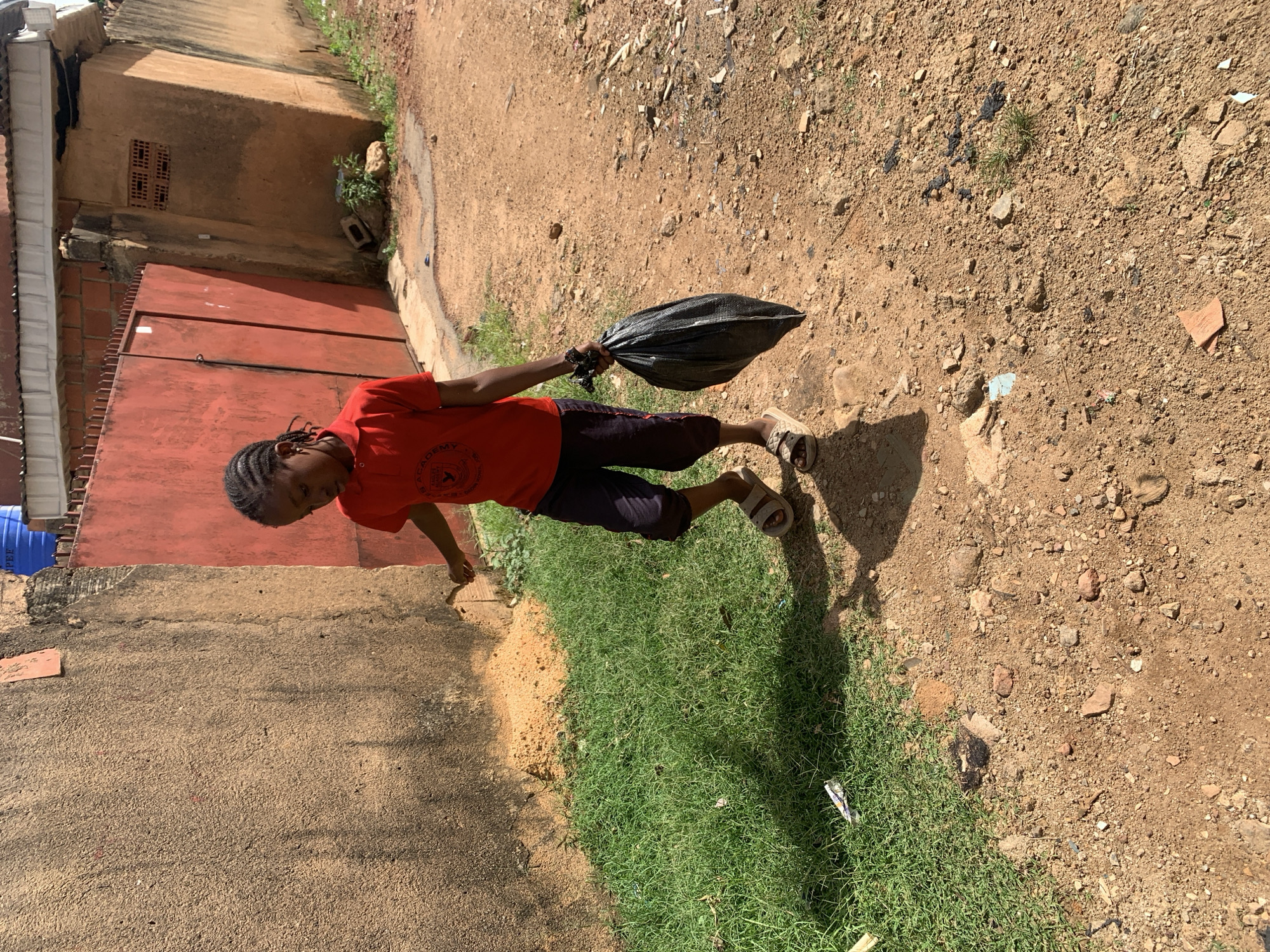 A young girl wearing a red t-shirt and dark shorts walks down a dirt path. She is carrying a black plastic bag in her right hand. The path is lined with sparse green grass on the left and a textured, light brown wall on the far left. Behind the girl is a large red metal gate. The ground is covered in loose dirt, small rocks, and scattered debris. The scene appears to be in an outdoor urban environment, possibly a residential area, with a building visible in the background. The lighting suggests it is daytime.