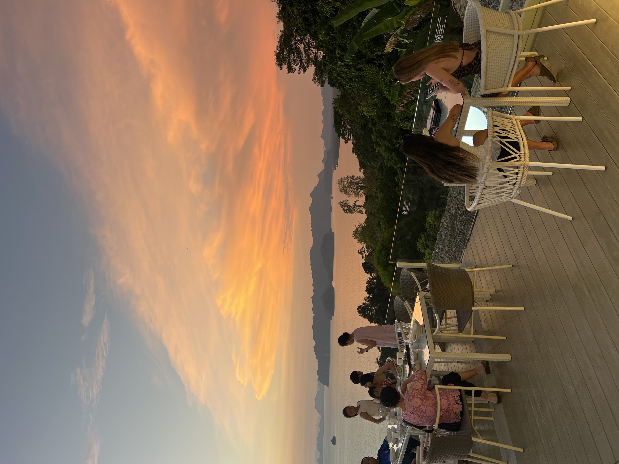 The image depicts a scene at a restaurant or outdoor dining area, likely in Khao Thong, Thailand, given the tropical vegetation and island landscape visible in the background. The sky is a vibrant mix of orange and light blue hues, suggesting sunset. Several tables are occupied by people who appear to be dining or socializing. Two women are seated at a white wicker table, one of whom is looking at a bright screen, likely a phone or tablet. Other patrons are visible at adjacent tables, with various objects such as plates and glasses indicating food service. The area is on a wooden deck or terrace with a glass railing. The scene conveys a relaxed, outdoor dining experience with a scenic view.