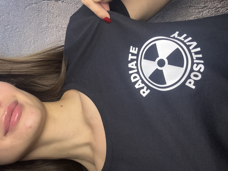A woman wears a black t-shirt with a silver design. The design includes a radiation symbol surrounded by the words "RADIATE" and "POSITIVITY." Her light brown hair is visible near her left shoulder. A manicured hand with red nail polish rests on the t-shirt's shoulder. Her face, including glossy lips, is partially in view. The background appears to be a textured grey surface.