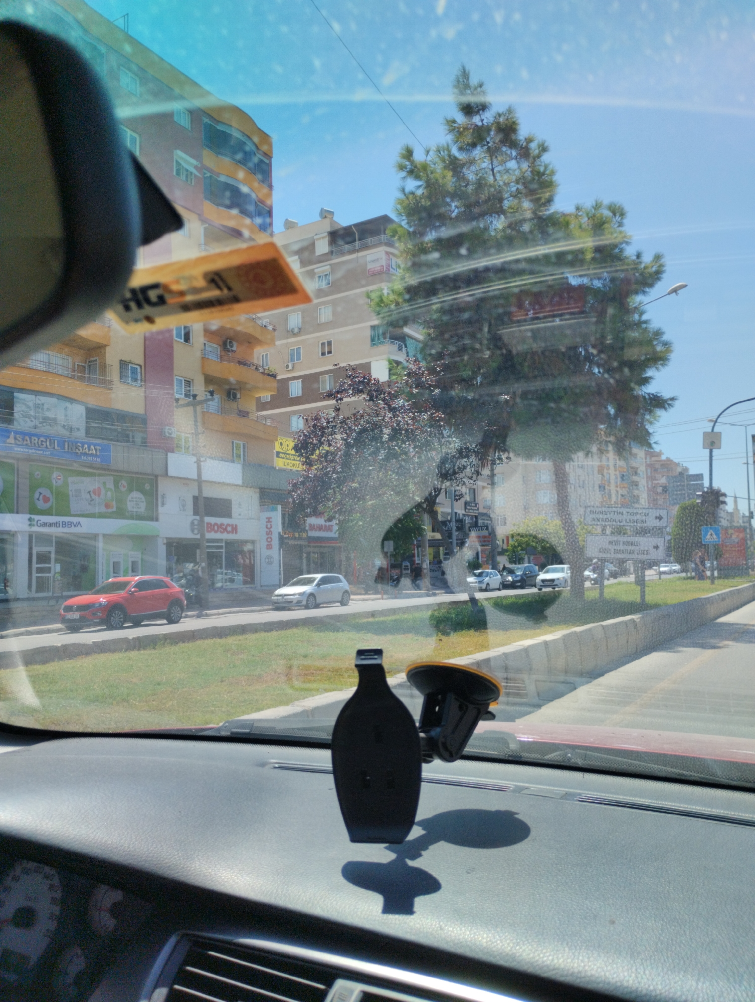 The image shows a view from inside a car, looking out through the windshield at a street scene in Mezitli, Turkey. A black phone mount with a suction cup is visible on the dashboard. The street is lined with buildings, including apartments and shops. Signs for businesses like "Sargül İnşaat" and "Garanti BBVA" are visible. A red SUV and a white hatchback are parked on the street, and several other vehicles are in motion. Trees and a sidewalk are also present, and a road sign indicates directions to "MEZİTLİ TOPCU ANADOLU LİSESİ" and "MEZİT KOLLARI GİZEM BAHAR LİSESİ". The sky is clear and blue, suggesting a sunny day.