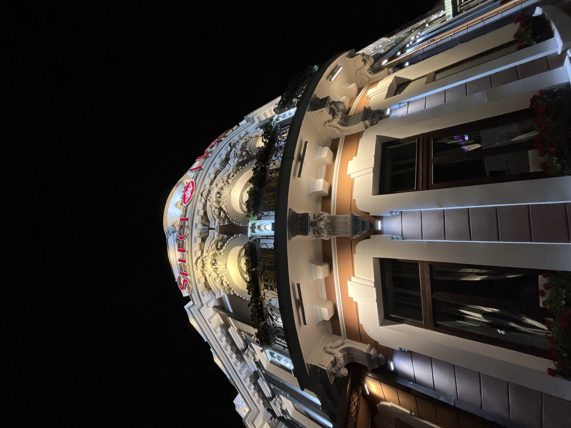 The image shows the exterior of a building at night, with the red neon sign "SELECT" illuminated at the top. The building appears to be ornate, with detailed architectural features such as columns, moldings, and a balcony with wrought-iron railings. The windows of the building are dark, suggesting they are closed or the interior lights are off. There are planters with red flowers on the balcony and below some of the windows. The building is located in the city of Iași, Romania, likely on a street with other buildings and some street lighting visible in the distance on the right side of the frame. The perspective is from a low angle, looking up at the building against a black night sky. There are no people visible in the image.