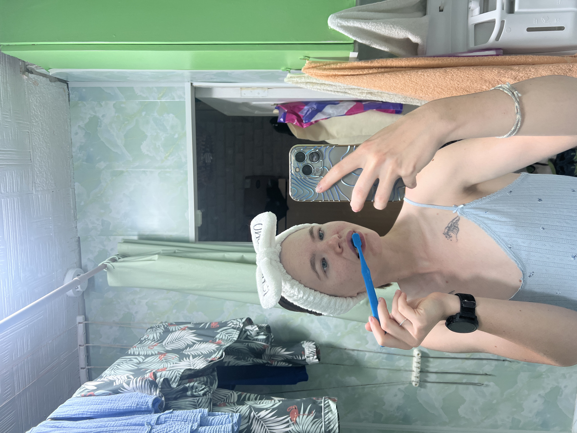 A young person is brushing their teeth in a bathroom. The person is wearing a light blue tank top, a white headband with a bow, and a black watch. They are holding a blue toothbrush in their right hand and are looking at their reflection in the mirror. The bathroom walls are covered in a light green marble-patterned material. There are clothes hanging on a rack to the left of the person, and towels hanging on the wall to the right. The ceiling appears to be made of textured panels. The image is a selfie taken with a smartphone. The specific location of Chaikovsky, Russia is not evident from the provided image.