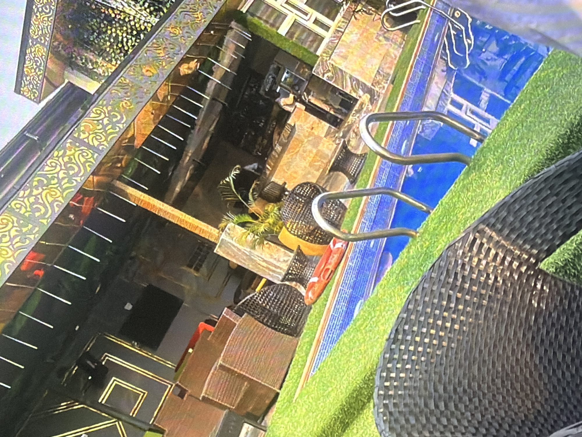 The image captures a poolside area in a city location identified as Bukuru, Nigeria. In the foreground, a portion of a dark woven chair is visible. Beyond the chair, a swimming pool with blue water and metal ladder rails occupies the mid-ground. The pool is bordered by a bright green artificial turf surface. In the background, a building with an ornate, patterned roof is illuminated. Part of the building's facade appears to be a bar area, with several individuals visible behind the counter. Various pieces of outdoor furniture, including woven chairs and lounge chairs, are scattered around the area. One red inflatable object rests on the artificial turf near the pool's edge. The overall scene suggests an outdoor recreational or entertainment venue.