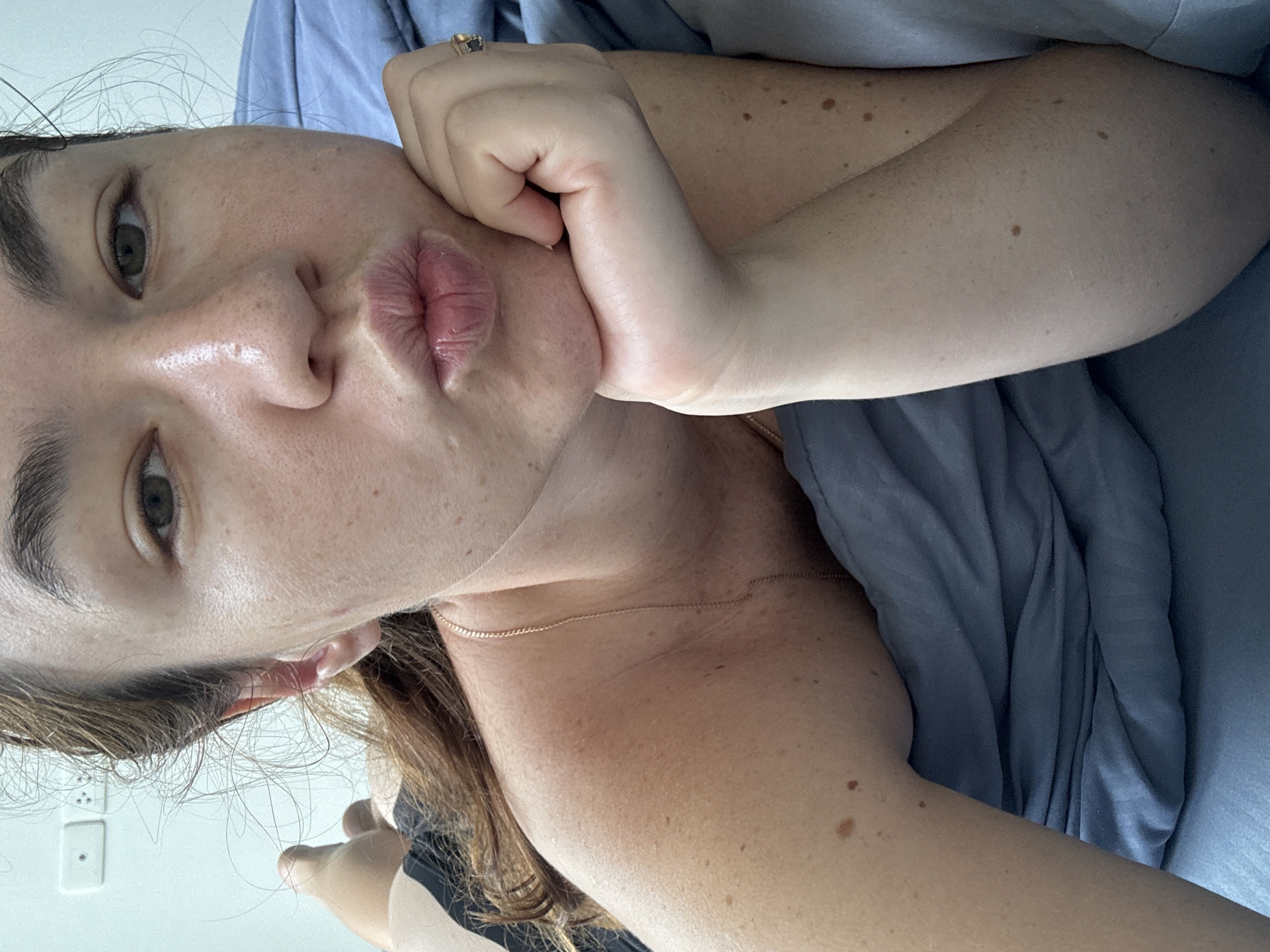 A close-up, overhead shot captures a woman lying on her side in bed, with her left arm bent and resting her chin on her hand. She is looking towards the camera with her lips pursed, as if blowing a kiss. Her hair, a shade of brown, is styled in a messy bun, with strands falling around her face and neck. She wears a gold chain necklace around her neck. She is wearing a black garment with a pattern on her shoulder. The background consists of a light blue duvet and a white wall with two electrical outlets. The woman's skin is visible, showing several freckles. The scene appears to be within a bedroom in Ratsada, Thailand, evidenced by the electrical outlets.