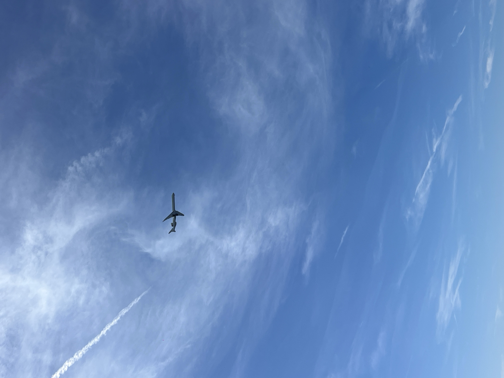 An airplane is flying across a bright blue sky with wispy white clouds. The airplane is silhouetted against the sky and appears to be a commercial jet. There is a long, thin contrail extending from the airplane. The image is taken from below, looking up at the sky. There is no visible indication of the city of Nice, France, or any other specific location.