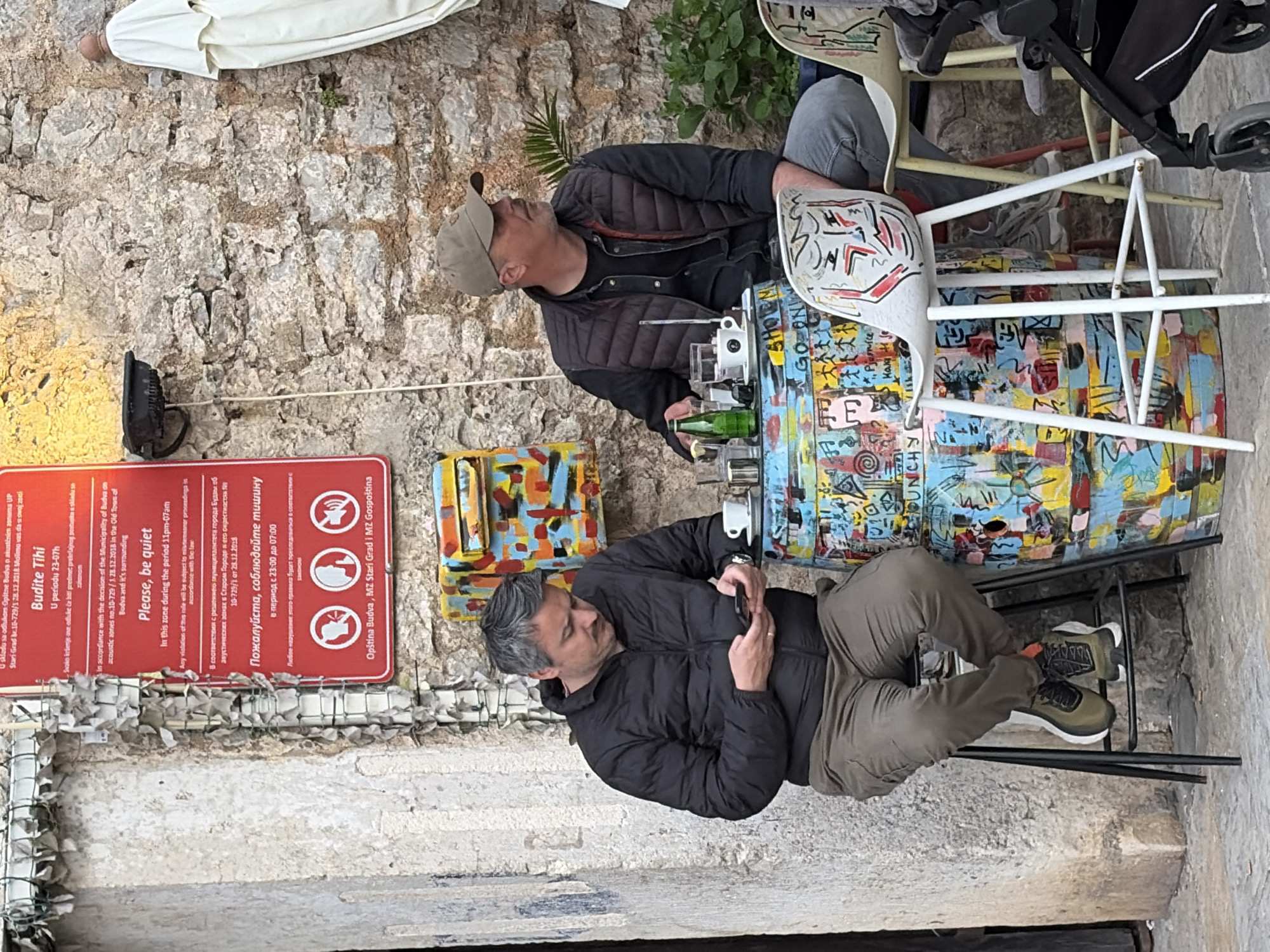 The image depicts a scene in Budva, Montenegro, possibly in the Old Town area, as indicated by a sign concerning noise restrictions ("Budite Tihi," "Please, be quiet"). Two men are seated at a colorful, art-covered barrel repurposed as a table. One man, wearing a brown baseball cap, dark quilted jacket, and light-colored pants, gazes upward. The second man, with graying hair and a dark quilted jacket, looks at his mobile phone. The table holds glassware and bottles. The location appears to be an outdoor space along a stone wall. A sign about noise restrictions is present on the wall. There is an empty stroller present.