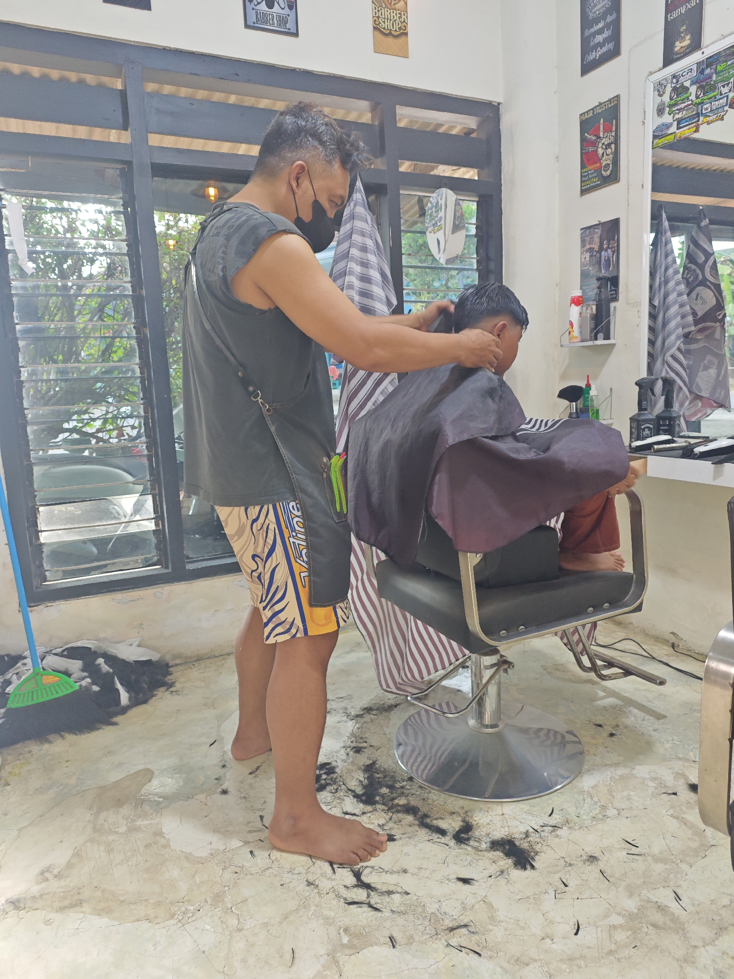 In a barbershop in Sarongan, Indonesia, a barber wearing a black face mask and a dark grey sleeveless shirt is giving a haircut to a young boy. The barber is standing and using electric clippers on the back of the boy's head. The boy is seated in a black barber chair with a red and white striped cape draped around him. He is wearing brown pants. The floor of the barbershop is made of light-colored tiles and is littered with clippings of dark hair. A green broom is leaning against the wall to the left of the scene. In the background, there are windows with slatted blinds, and shelves on the wall display various bottles and tools. Several framed pictures and posters are also visible on the walls. The overall impression is of a functional, everyday barbershop environment.