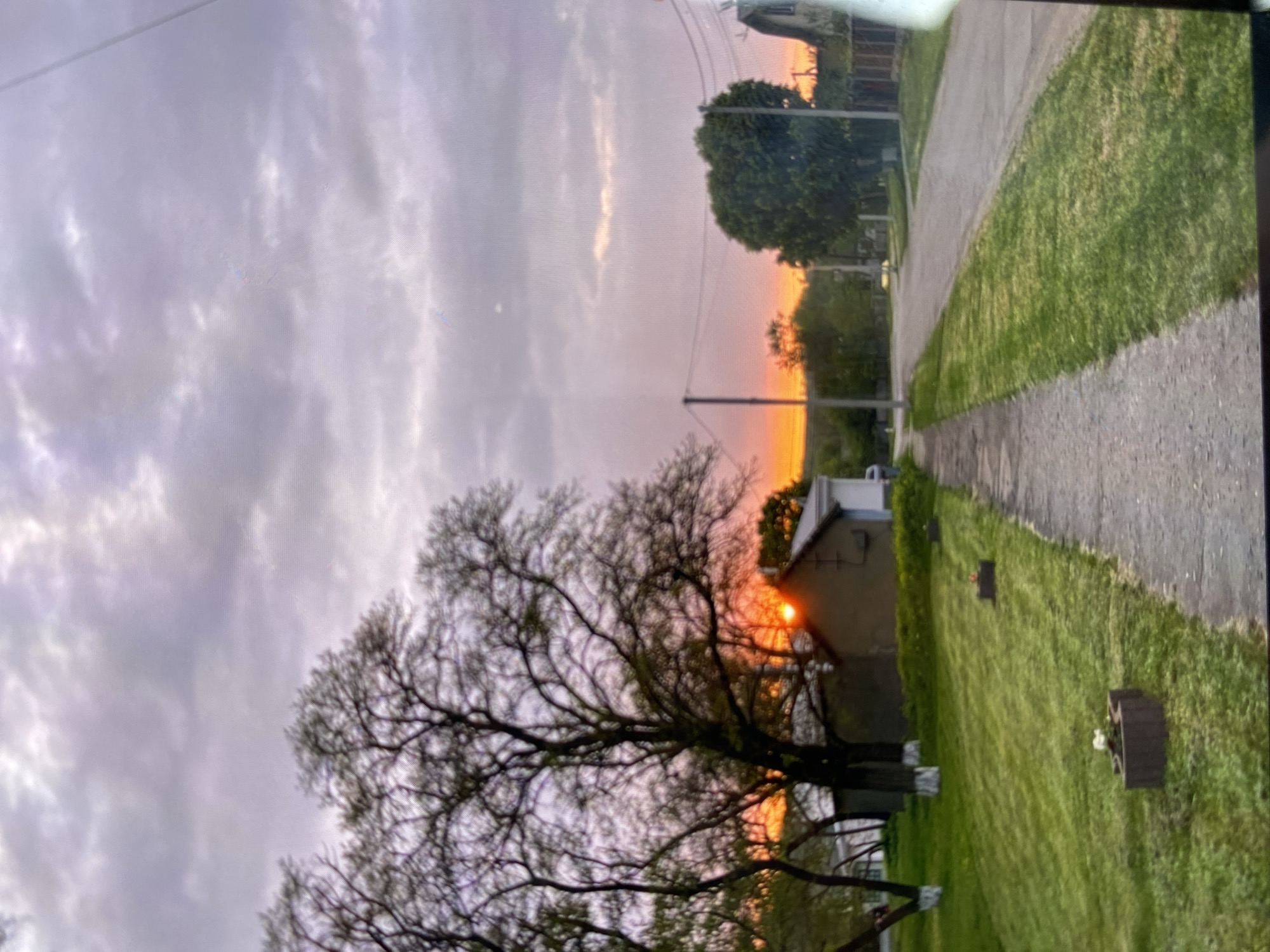 The image depicts a rural street scene during a sunset. The sun is visible on the horizon, casting a warm orange glow that is reflected in the clouds. The sky is a mix of purple and orange hues. In the foreground, a paved sidewalk runs along the right side of the image, bordered by a strip of well-maintained green grass. Beyond the grass, several trees with bare branches are scattered across the landscape.
On the left side of the scene, a small building with a dark roof and light-colored walls is partially visible, nestled amongst the trees. The building appears to be a house or a shed. Further down the street, a few more houses and trees can be seen. The street itself is paved and appears to be in good condition. A few utility poles and wires are visible along the right side of the street. The overall impression is one of a quiet, peaceful evening in a rural setting.