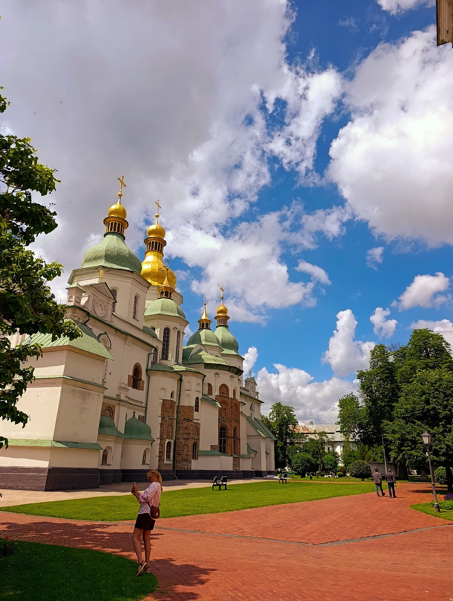 The image depicts the exterior of a magnificent church with multiple green and gold domes topped with crosses, indicative of Orthodox Christian architecture. The building is large and imposing, with white walls and brick accents. It is set within a well-maintained park-like area featuring lush green lawns and mature trees. A paved red brick pathway winds through the scene.
In the foreground, a woman with blonde hair is standing on the pathway, holding a smartphone up as if taking a photo or video. She is dressed casually in a light-colored shirt and dark shorts, with a small bag slung over her shoulder. Further in the background, two men are walking away from the camera on the red brick path.
The scene is bathed in bright sunlight, suggesting it is daytime, likely late morning or afternoon. The sky is a vibrant blue with scattered white, puffy clouds. The overall atmosphere appears peaceful and picturesque, hinting at a pleasant day for sightseeing or visiting a historical landmark. The architecture and the presence of the church strongly suggest the location is within Kyiv, Ukraine, a city known for its many religious buildings. No visible text is present in the image.