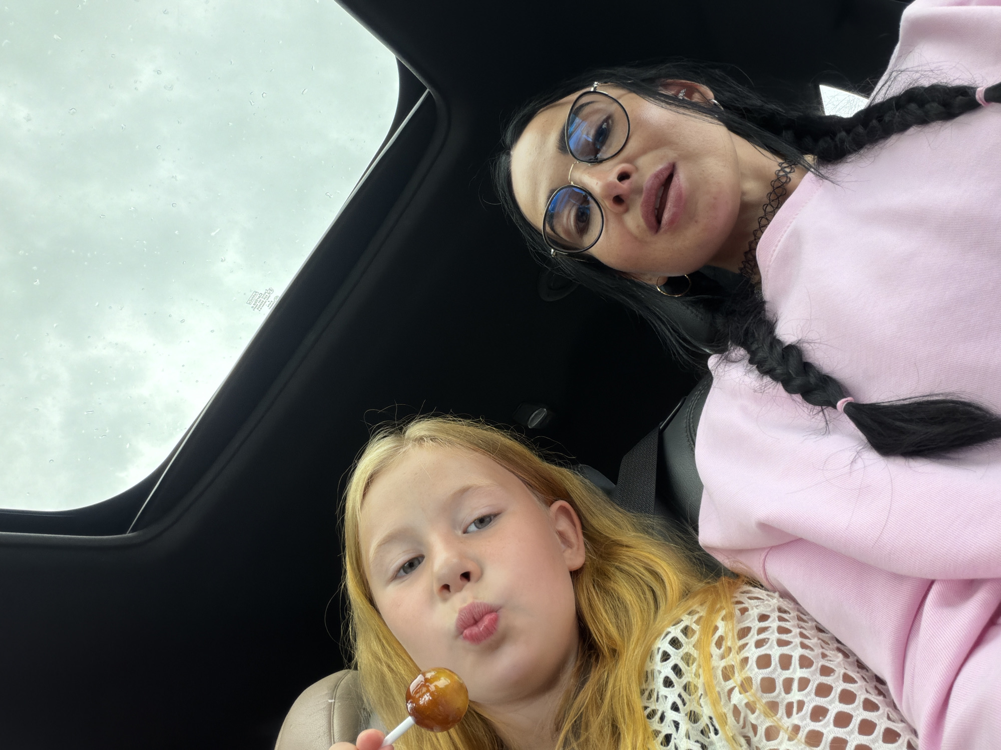 Two people are inside a car with a sunroof open, looking up at a cloudy sky. On the left, a young girl with long blonde hair is holding a lollipop and puckering her lips. She is wearing a white mesh top. On the right, a woman with dark hair in braids and wearing glasses and a pink sweatshirt is looking forward. A black choker necklace is visible around her neck. The car's interior is visible, including the sunroof frame and black upholstery. The location is specified as Sholokovo, Russia.
