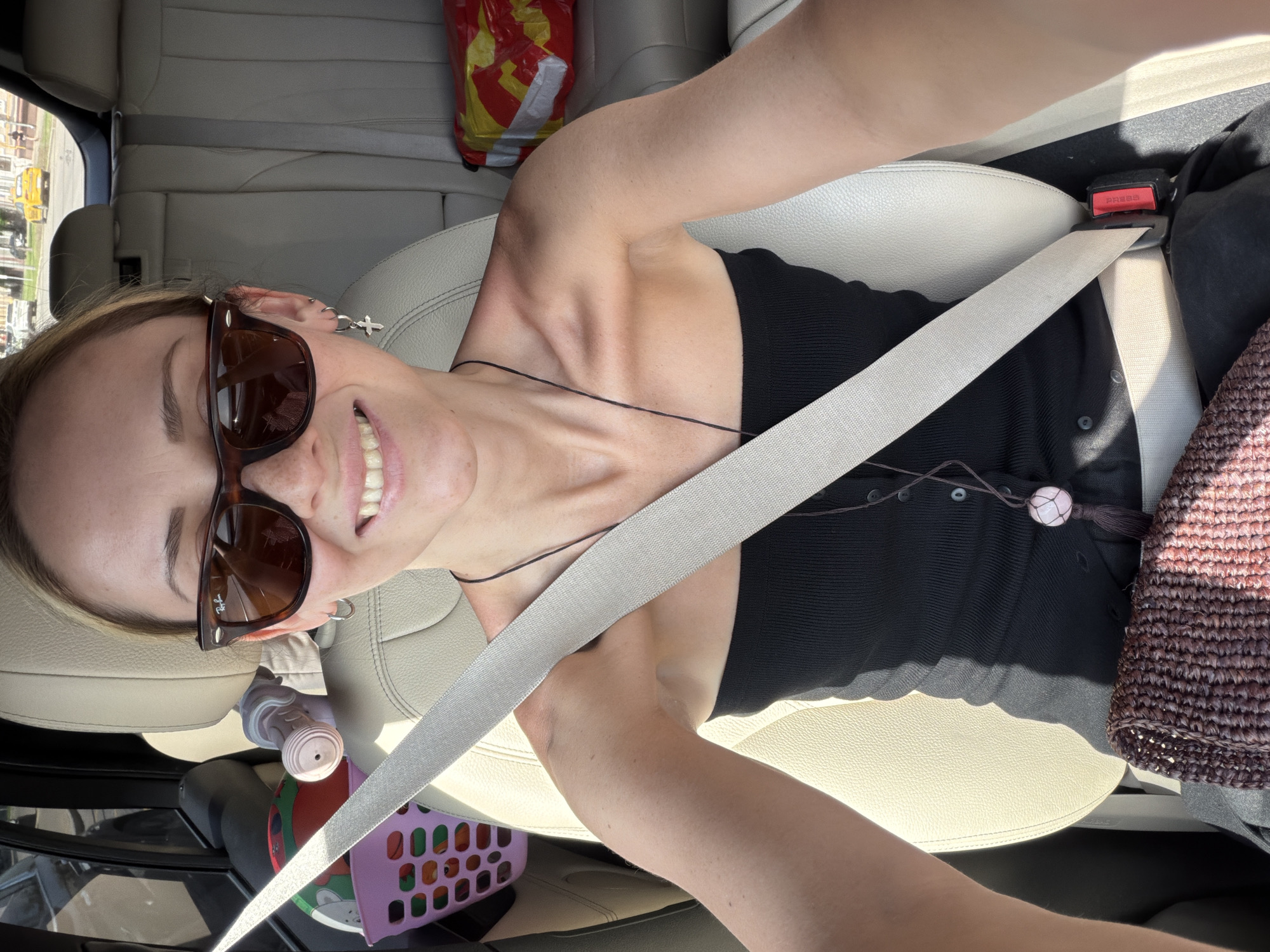 The image depicts an adult female taking a selfie from the front passenger seat of a car on a sunny day in Moscow, Russia. The fair-skinned female has blonde hair, visible freckles, and is smiling with her teeth exposed. She is wearing dark brown Wayfarer-style Ray-Ban sunglasses, a black strapless top, and a thin dark cord necklace featuring a light pink spherical pendant and a dark tassel. She wears a silver hoop earring with a small cross charm on her right ear and a plain silver hoop on her left ear. A light-colored seatbelt is fastened across her chest and lap, secured by a red buckle labeled "PRESS". The car's interior features light-colored seats. In the rear left seating area, a pink plastic basket contains a multi-colored ball and a cylindrical object. On the rear right seat, a red and yellow plastic bag is visible. Through the car's rear window, a street scene is observable, including buildings, other vehicles, and a yellow car in the distance. Bright sunlight casts distinct shadows within the car's interior.
