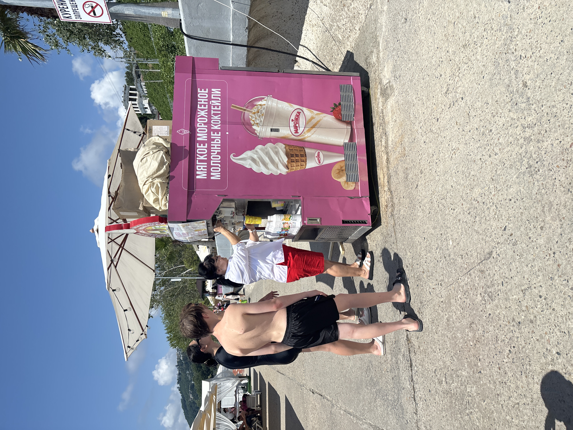 An outdoor, ground-level, eye-level, medium shot captures a scene on a paved walkway near the Black Sea in Sochi, Russia. The bright, sunny day illuminates a pink, wheeled ice cream and milkshake stand. The stand features graphics of a swirl ice cream cone and a milkshake in a cup, along with the Russian text "МЯГКОЕ МОРОЖЕНОЕ МОЛОЧНЫЕ КОКТЕЙЛИ" (Soft Ice Cream, Milkshakes). A woman in a white shirt and red shorts is operating the stand, attending to a customer. In the foreground, three individuals are visible. A young man, shirtless and wearing black swim shorts, stands in the center. To his left, a woman in a black swimsuit looks towards the stand. Another woman, partially obscured, stands to the right of the young man. In the background, white beach umbrellas and lounge chairs suggest a resort or beach area. A white building with a distinctive architectural style is visible on a lush green hillside, characteristic of the Sochi region. A sign with a red circle and slash over a cigarette indicates a no-smoking zone.