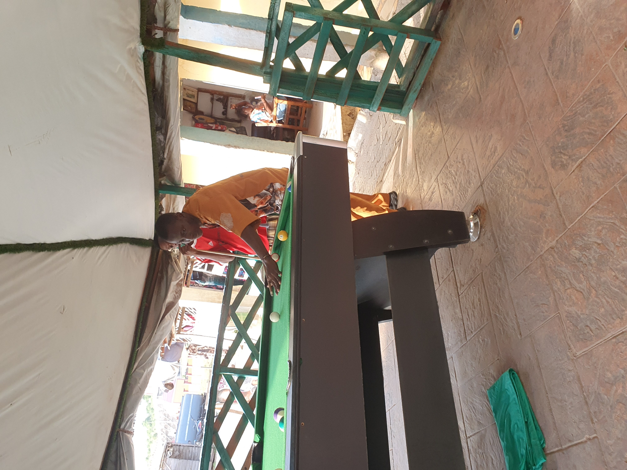 In Lokoja, Nigeria, two men are playing billiards under a white canopy. One man, wearing an orange shirt and brown pants, is actively engaged in the game, holding a cue stick and aiming at a ball on the green felt-covered pool table. Another man, partially visible in a red shirt, stands nearby. The scene takes place on a paved outdoor patio, with a green wooden railing framing the billiard area. In the background, a woman sits on a chair inside a building. The environment appears to be a casual recreational space, possibly a bar or outdoor lounge, with various objects visible in the background, including shelves with items and a motorcycle parked outside.