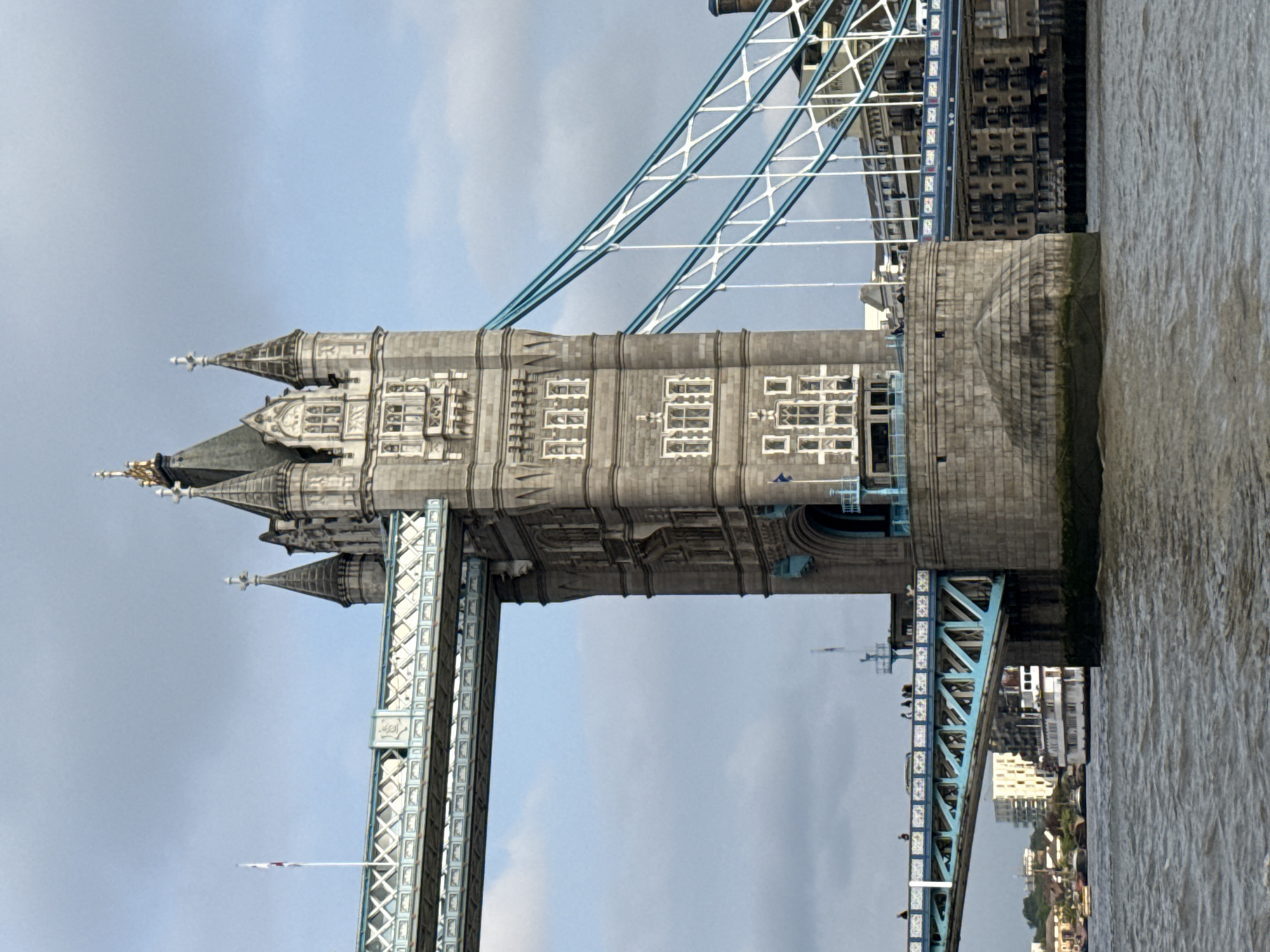 The image features a segment of the Tower Bridge in London, United Kingdom. The bridge's stone tower is prominently displayed, adorned with multiple windows and gothic-style spires. Steel cables connect the tower to the upper structure of the bridge. A part of the Thames River is visible to the right of the bridge. The sky is overcast. Other buildings and structures are partially visible in the background.