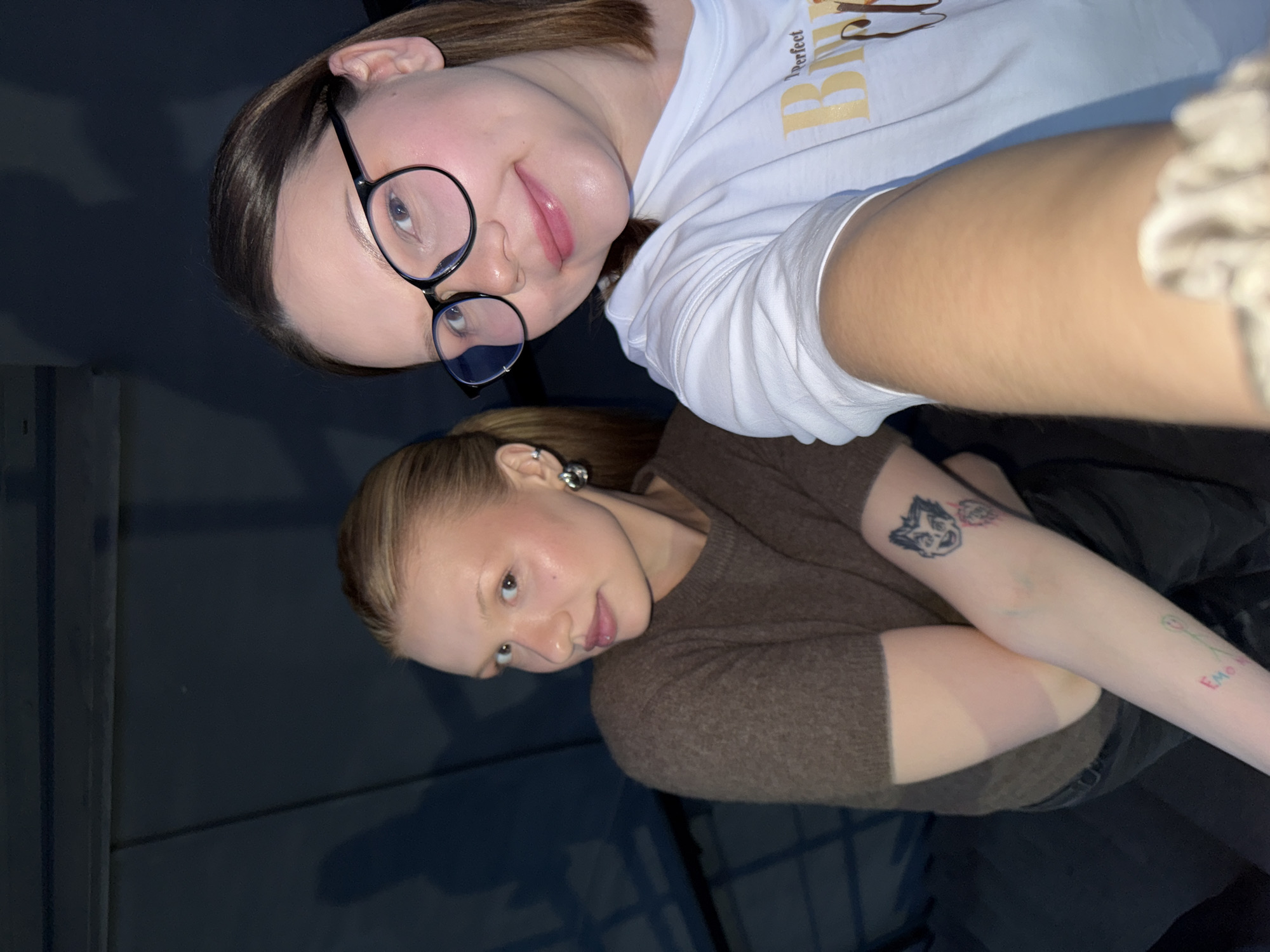 The image is a selfie of two young women. One woman is wearing glasses and a white T-shirt with the word "Perfect" printed on it. The other woman is wearing a brown sweater and has tattoos on her arm. The background is dark. The image was taken inside in the city of Chaikovsky, Russia, with the location being a bedroom.