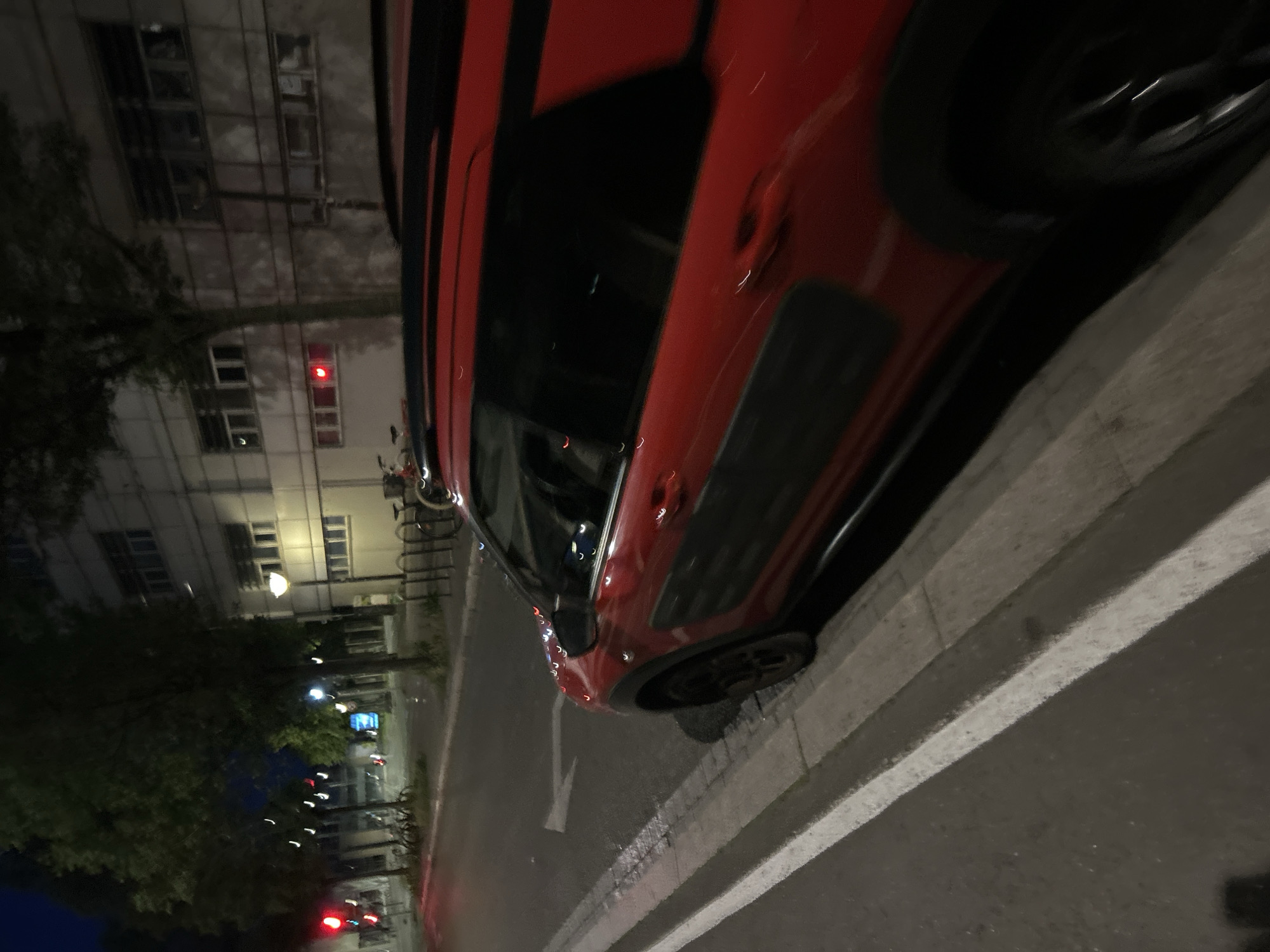 The image captures a nighttime street scene in Strasbourg, France. A red Citroën C4 Cactus, identified by its black side panels, is parked parallel to a concrete curb on the right side of an asphalt road. The vehicle's front right wheel is positioned near the curb. The road surface displays a solid white line parallel to the curb and a white directional arrow pointing left. Adjacent to the road is a narrow sidewalk constructed from grey paving stones.
To the left, a line of mature trees with dark foliage borders the street, partially obscuring the dark night sky. Multiple streetlights and a red traffic signal are visible further down the road. Several bicycle racks are present on the sidewalk, some containing bicycles. On the right, beyond the parked car, stands a multi-story building with a light-colored facade featuring numerous rectangular windows. A prominent streetlamp with a warm yellow glow illuminates the building's exterior. A red rectangular light fixture is affixed to the building's lower section. No individuals are present in the scene.
