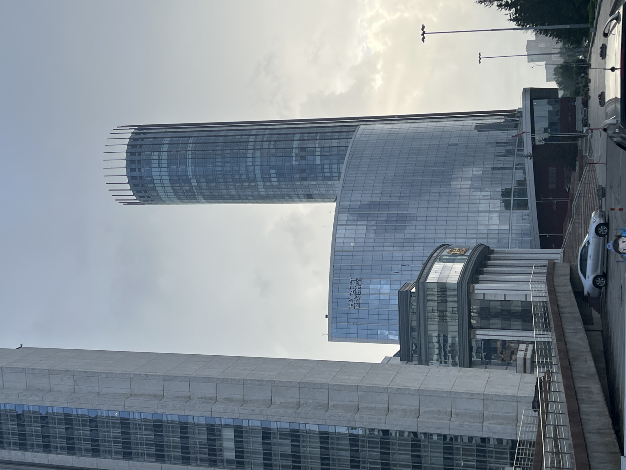 The image displays an urban scene in Yekaterinburg, Russia, featuring prominent modern architecture under an overcast sky.
In the center-right of the frame stands the Iset Tower, a tall, cylindrical skyscraper with a blue-tinted glass facade. Its apex is distinctive, featuring a crown of evenly spaced, dark vertical elements. Adjacent to and below the Iset Tower is the Hyatt Regency Yekaterinburg, a large building with a curved, blue-tinted glass facade that reflects the cloudy sky. The hotel's name, "HYATT REGENCY," is prominently displayed on its upper section. Below the Hyatt Regency, a lower structure is visible, characterized by a series of vertical columns and a gold-colored emblem, likely part of the complex.
On the far left, a tall, multi-story building with a light-colored, possibly concrete or stone, facade occupies a significant portion of the left edge. This building features vertical sections of darker, recessed windows.
In the mid-ground and foreground, a network of concrete ramps with metal railings ascends towards the buildings. A silver sedan, likely a Toyota Camry, is parked on one of these ramps. Part of a darker vehicle is visible in the extreme bottom right corner of the image. Streetlights with multiple fixtures are positioned on the right side of the frame. Beyond them, a line of coniferous trees and other distant buildings are visible against the hazy, cloudy sky. The overall lighting suggests a cloudy daylight condition, with soft, diffused light.