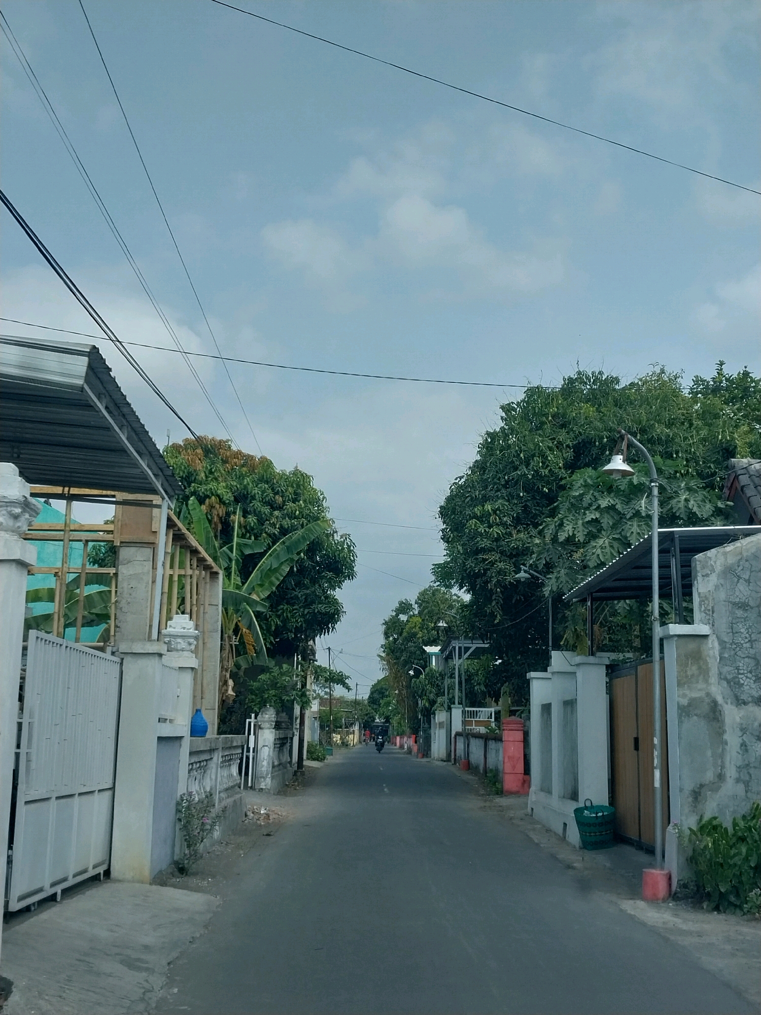 The image depicts a residential street in Purwomartani, Indonesia, under an overcast sky. A paved road runs through the center of the frame, flanked by houses and fences. On the left, a white metal gate is prominent, adjacent to a white concrete fence with decorative elements. Beyond the fence, a building under construction is visible, with visible wooden framing and blue window frames. Lush green trees, including banana trees, line the street, with some showing signs of seasonal color change. On the right side of the road, a concrete wall with textured surfaces and a wooden gate are visible. A green woven trash bin rests near the entrance. A street lamp with a distinctive arched arm is mounted on a pole. Further down the street, another street lamp is visible. In the distance, a person on a motorcycle is traveling away from the camera. Power lines crisscross the sky above. The overall scene suggests a typical day in a suburban or semi-urban area in Indonesia.