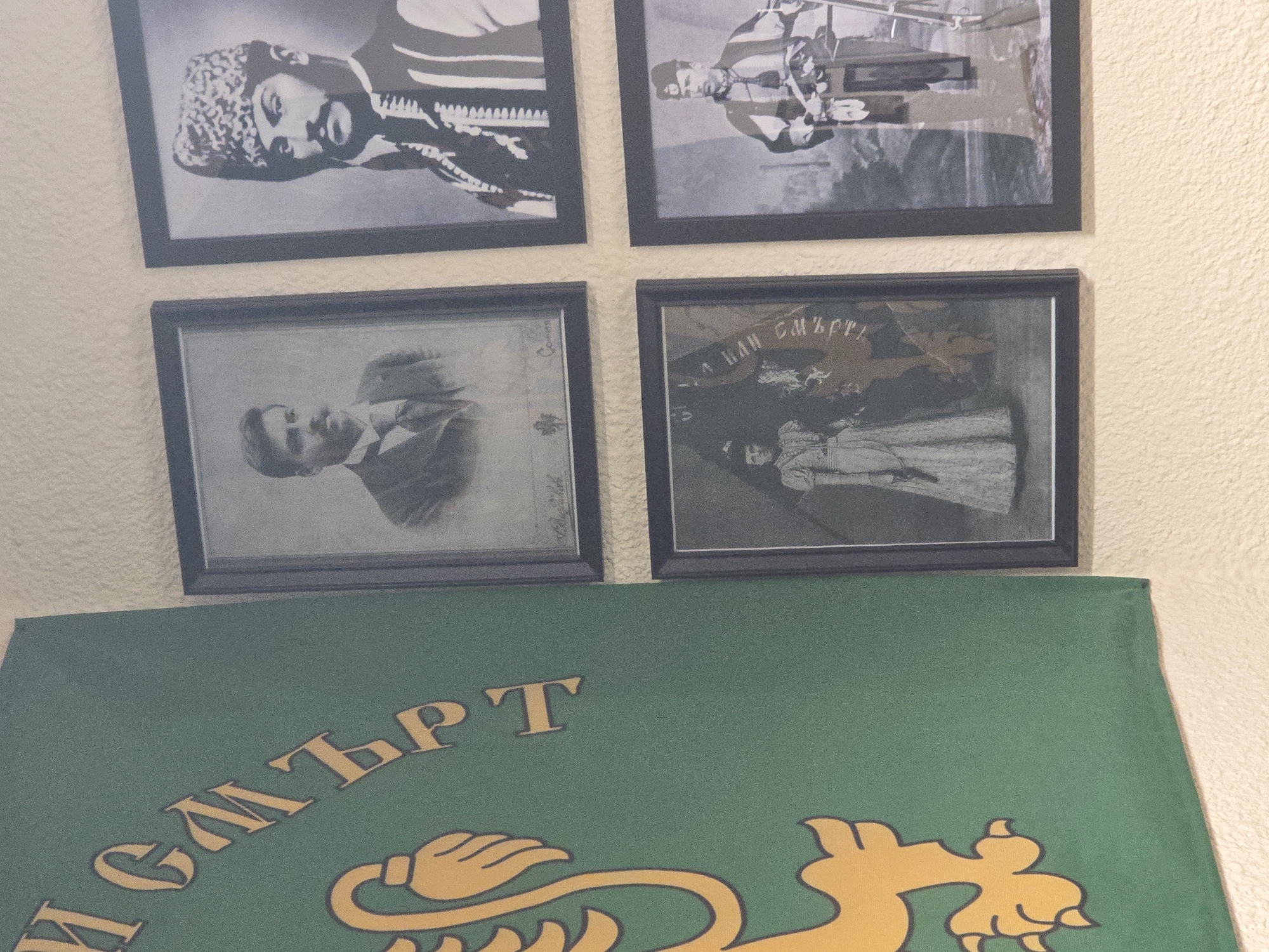 A close-up shows four framed black and white photographs hanging on a light beige textured wall above a green flag with yellow writing and a yellow lion emblem. The photographs are arranged in a square configuration, two above and two below. The top left photograph depicts a man wearing a patterned hat and a traditional garment. The top right photograph shows two men standing near a mechanical device. The bottom left photograph shows a man in a suit and tie. The bottom right photograph shows a group of people standing with a banner that has text in a foreign language. The green flag below has text in a foreign language and a yellow lion emblem. The location appears to be indoors on a wall.