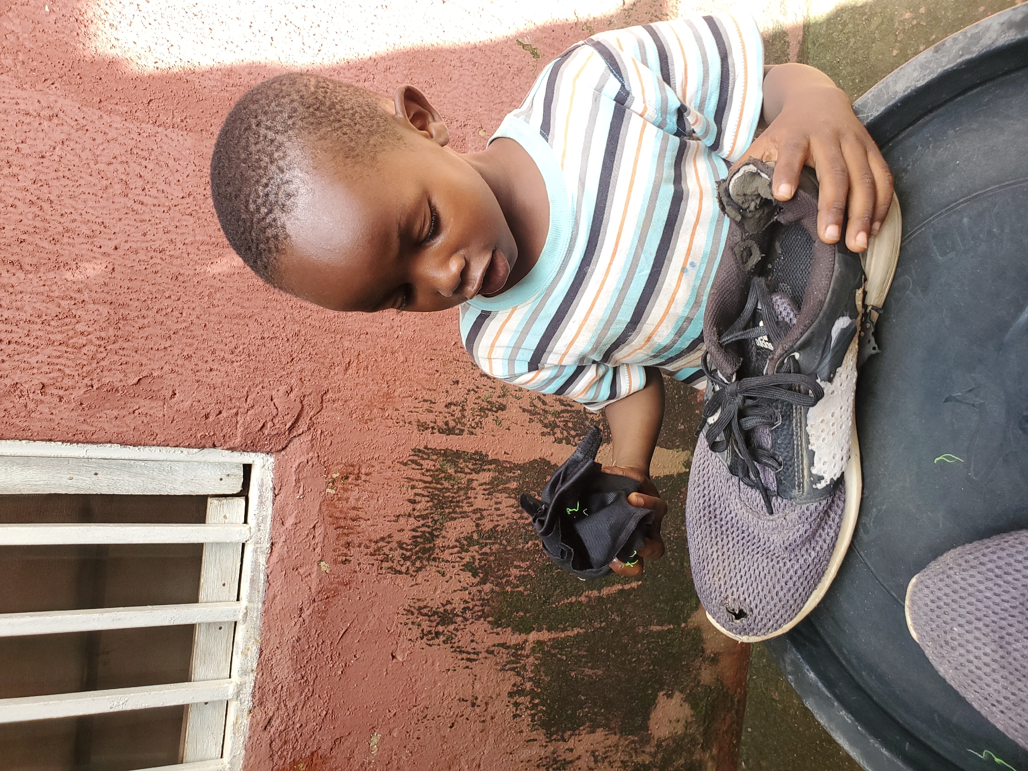 A young Black boy, likely between 3 and 5 years old, is seated outdoors, holding a torn black shoe. He is wearing a striped t-shirt with horizontal bands of light blue, white, orange, and dark blue. His gaze is directed downwards towards the shoe. The shoe, which is grey and black with white accents, appears heavily worn and damaged. The boy is positioned in front of a reddish-brown, textured wall that shows signs of weathering and some patches of green growth. To the left of the frame, a window with white vertical bars and a wooden frame is visible against the wall. The ground appears to be a large, dark, circular object, possibly a basin or tub. The overall setting suggests a casual, possibly impoverished, urban environment in Jos, Nigeria. The lighting indicates it is daytime, with sunlight casting shadows on the wall.
