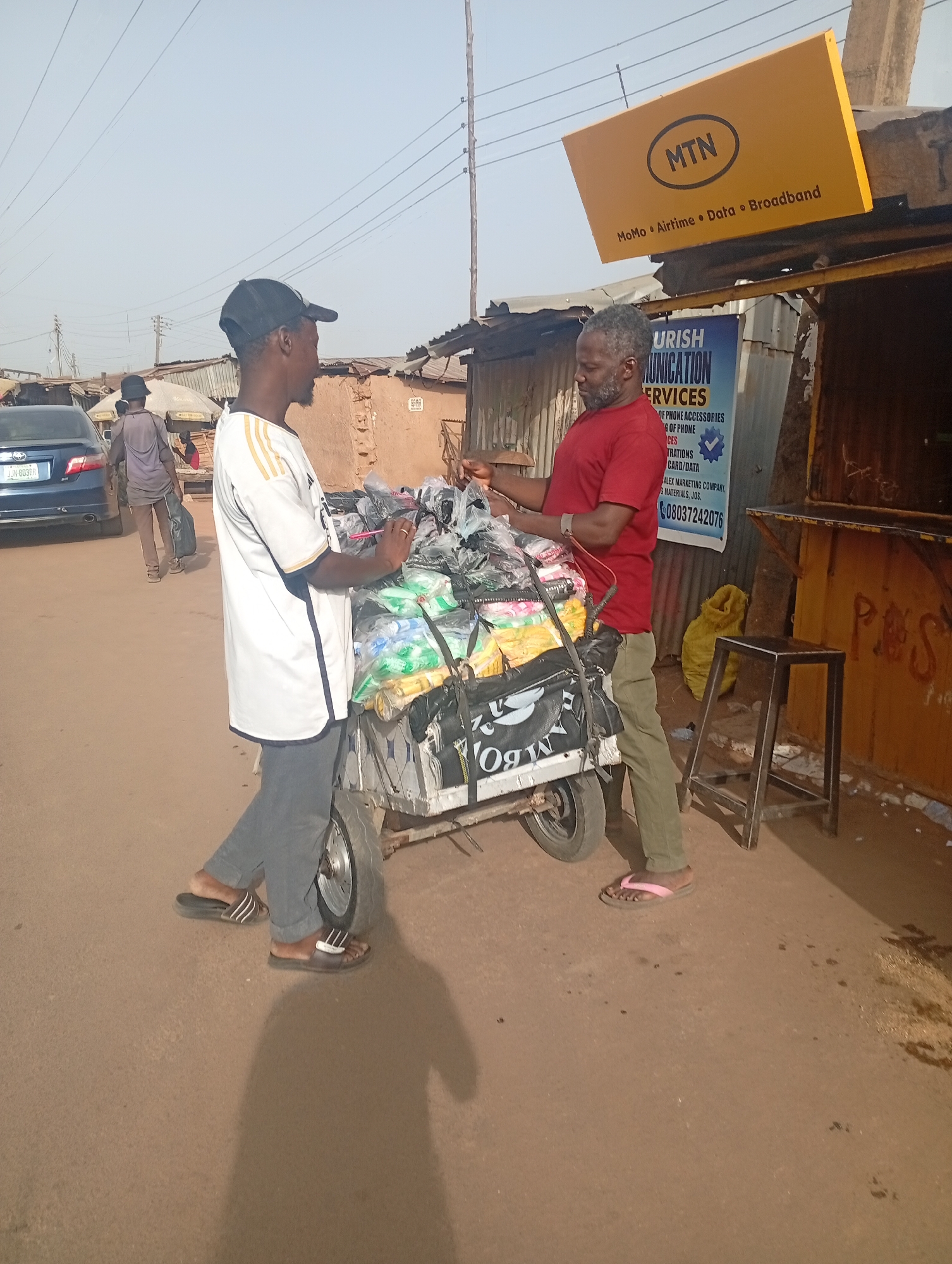 Two men are engaged in a transaction on a street in Jos, Nigeria. One man, wearing a red shirt and green pants, stands next to a roadside stall with a yellow MTN sign above it. He appears to be examining goods from a three-wheeled cart. The cart is loaded with what appear to be packaged items. The second man, wearing a white and black shirt and grey pants, stands next to the cart, seemingly selling the items. In the background, another person walks away from the scene, and a blue car is parked on the side of the street. The environment appears to be a busy marketplace or commercial area. A sign for "URISH COMMUNICATION SERVICES" advertising phone accessories and services is visible on the stall.