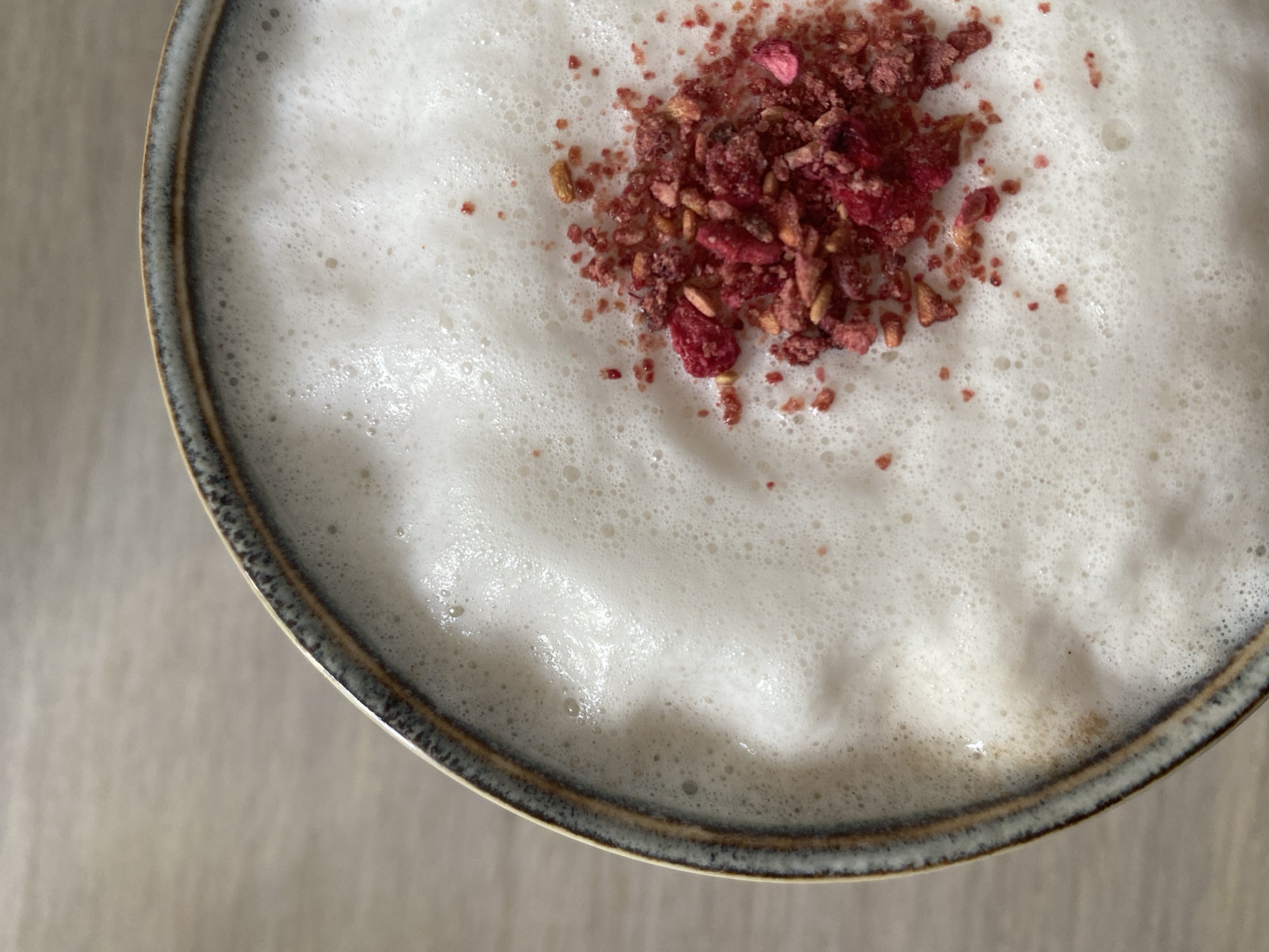An overhead, close-up shot depicts a partially filled cup containing a white, frothy liquid. The froth is topped with a scattering of dried, reddish-purple particles. The cup is ceramic with a grey-brown rim. The cup sits on a light-colored surface. There is no information in the image to indicate this photo was taken in Chaikovsky, Russia.