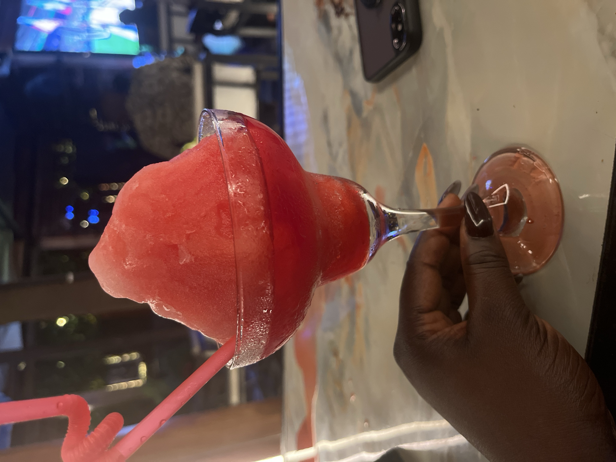 The image shows a close-up of a hand holding a frozen red cocktail in a margarita glass. Two pink straws are inserted into the drink. The drink is a vibrant red color and has a frozen, icy texture on top. The glass is clear and appears to be sweating, indicating it is cold. The hand holding the glass has dark skin and is wearing dark nail polish. In the background, out of focus, there are lights and what appears to be a screen showing a colorful image, possibly a television or projector. The setting looks like an indoor or covered outdoor area, with hints of ambient lighting suggesting a bar or restaurant environment. A portion of a table surface is visible in the foreground, with a marble-like pattern and what appears to be a cell phone in the right side of the frame. There is no specific indication of the city of Abuja, Nigeria.