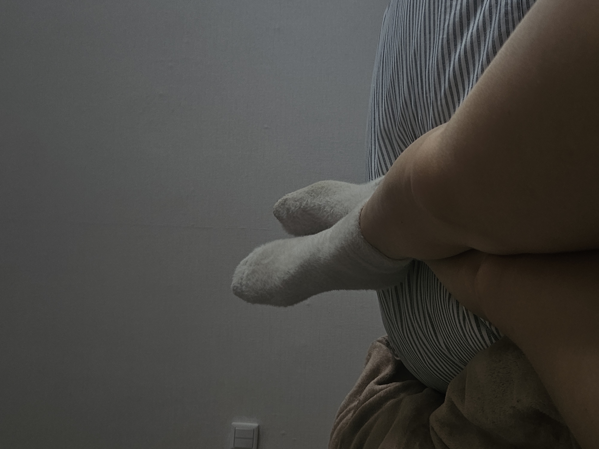 The image depicts a low-angle, close-up shot of a person's legs and feet resting on a bed. The person is wearing white, fuzzy socks, and their legs are crossed. They are lying on a pillow with blue and white stripes, covered by a brown blanket. The background shows a plain, light-colored wall with a light switch. The lighting is dim, suggesting it might be evening or a dimly lit room. There is no indication of the person's location in Chaikovsky, Russia. The scene appears to be indoors, in a bedroom.