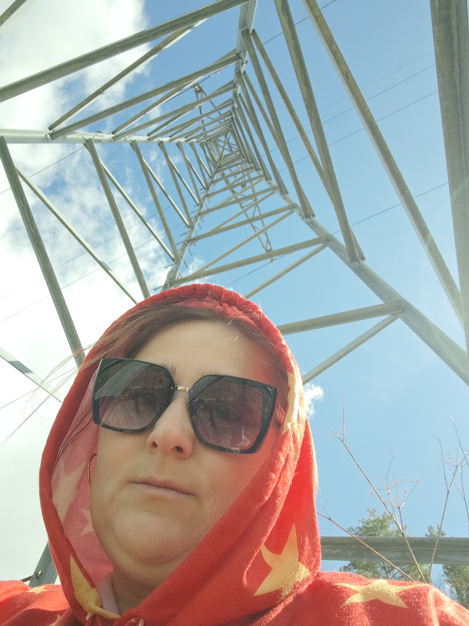 A low-angle, full-color photograph depicts a person wearing a red hooded sweatshirt with yellow stars and dark sunglasses, positioned in front of a large electrical tower. The tower is comprised of steel beams and spans upwards into the blue sky with scattered clouds. The person's face is central in the foreground. To the right side, there are thin, dried plant stalks. There's a hint of greenery and possibly a fence behind the person. The city of Chaikovsky, Russia, cannot be confirmed or denied based solely on the content of this image.