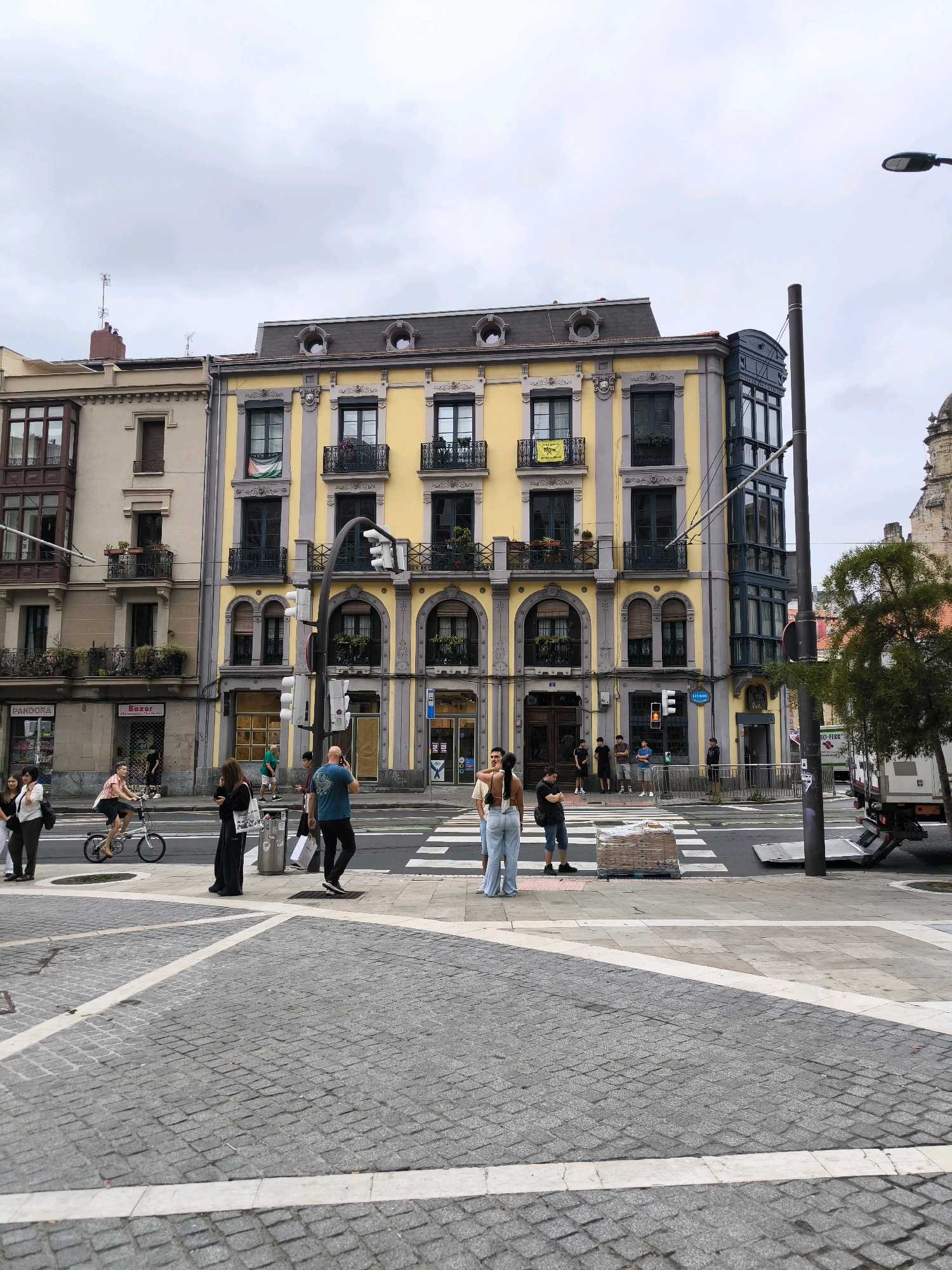 This image depicts a street scene in Bilbao, Spain, likely near the Ensanche district. The focus is on the intersection of streets with a prominent yellow building on the right, featuring ornate balconies and dark-paned windows. A pedestrian crossing is visible in the foreground, with several people waiting or walking across.
On the left, a lighter-colored building with bay windows stands next to the yellow structure. A woman on a bicycle rides across the crosswalk. Further into the scene, a delivery truck is parked on the right side. The sky is overcast, suggesting a cloudy day. The architecture and cobblestone streets are characteristic of many European cities, and the presence of Spanish signage further confirms the location.
Specific details include:
- **Buildings:** The yellow building has a distinctly European architectural style with decorative elements. The building to its left is lighter in color and has protruding bay windows.
- **People:** Individuals are engaged in various activities: crossing the street, waiting at the crosswalk, and cycling.
- **Objects:** A traffic light, a lamppost, a delivery truck, and street signs are visible. A pallet of goods is placed near the crosswalk.
- **Environment:** The ground is paved with cobblestones, and the sky is overcast. A tree is visible on the right side of the frame.
The overall impression is that of a typical urban street scene in a European city, with elements of daily life unfolding at an intersection.