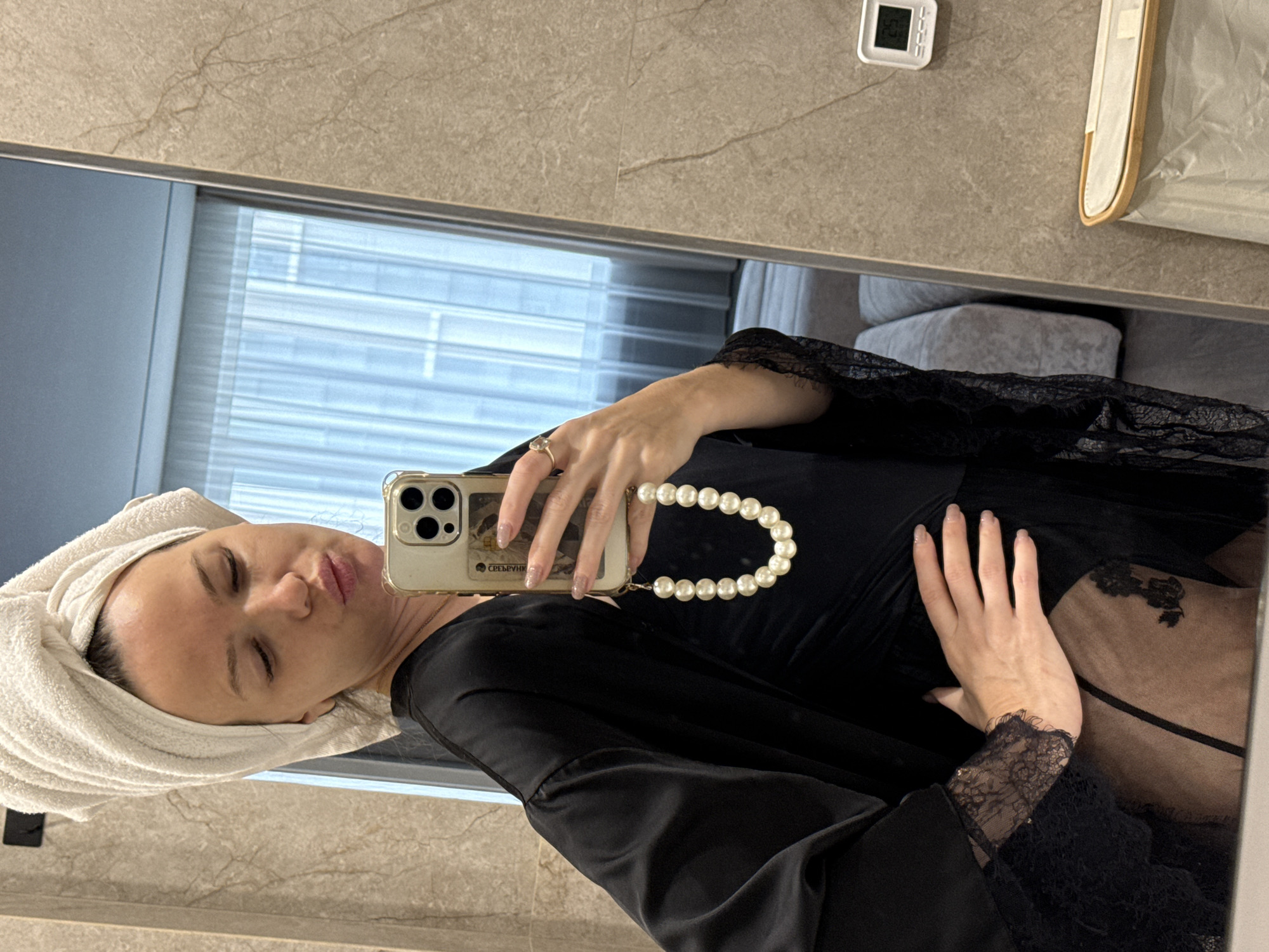 A woman is taking a selfie in a bathroom. She is wearing a black robe with lace trim and has a white towel wrapped around her head. She is holding a phone with a pearl wrist strap. The background shows a beige tiled wall and a window with blinds. The location is an unknown city and unknown.