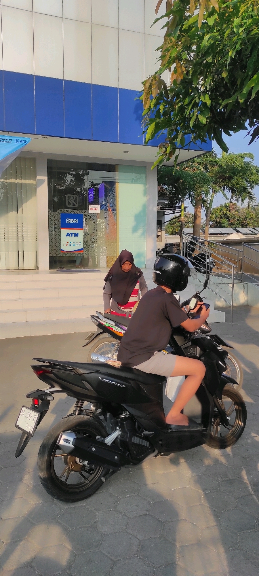 In Purwodadi, Indonesia, two individuals are near a BRI bank ATM. In the foreground, a person wearing a black helmet, a dark t-shirt, and light-colored shorts sits on a black scooter. They are holding a smartphone. Behind them, another person wearing a black hijab and a red and white striped shirt stands next to a motorcycle. The BRI bank features a blue and white facade with glass doors displaying ATM signage. The ground is paved with hexagonal stones, and trees are visible in the background. The lighting suggests it is either early morning or late afternoon.
