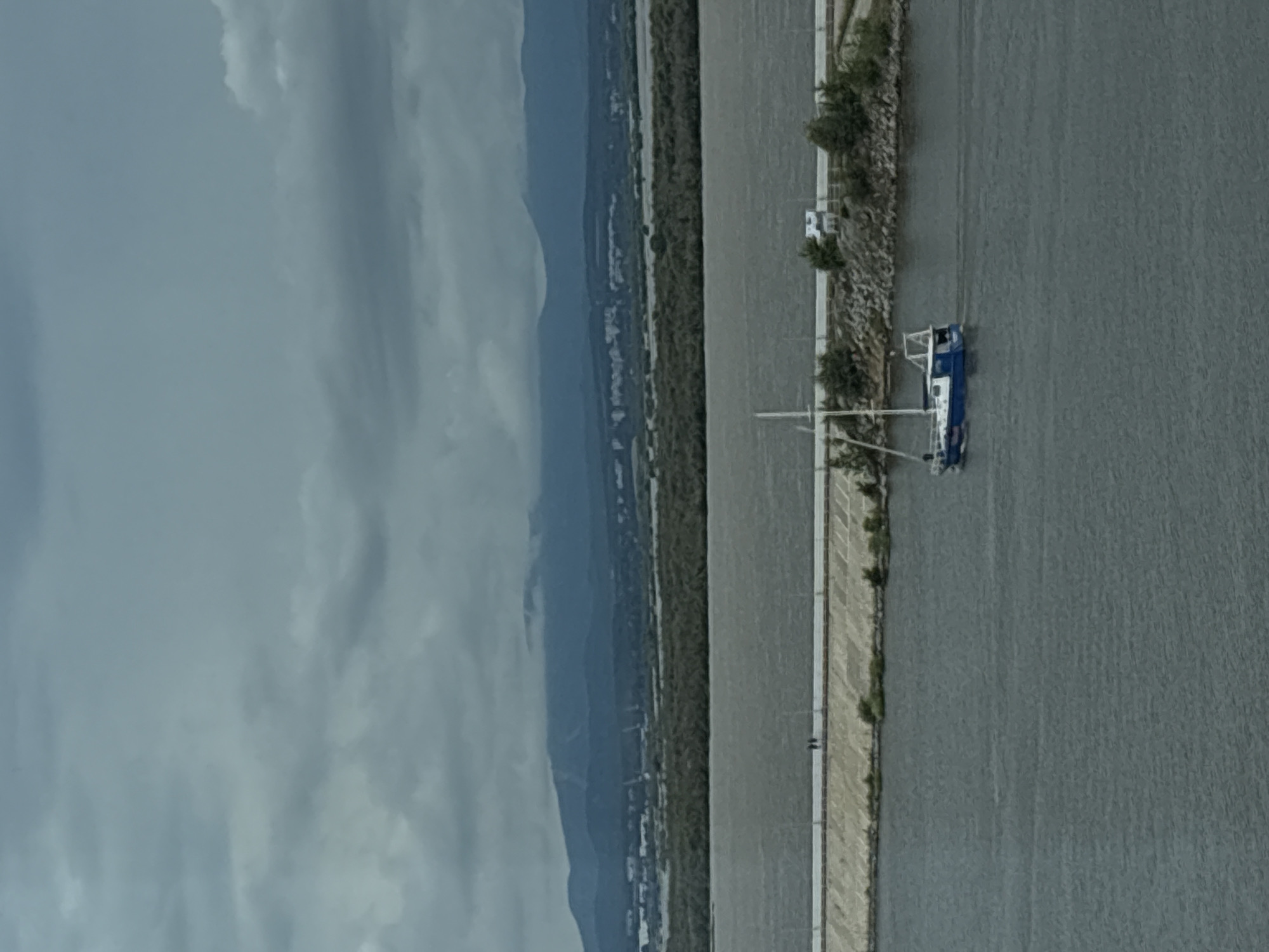 A blue and white sailboat navigates a wide, calm river under a heavily overcast sky. The river has a muddy, dark hue, and the sky is filled with thick, grey clouds, suggesting a potentially stormy or gloomy atmosphere. In the middle ground, a concrete embankment or levee runs horizontally across the image, separating the river from a strip of land. Two silhouetted figures are visible walking on the embankment. Beyond the embankment, a dense line of dark green trees stretches across the landscape. Further in the distance, a range of low, blue-grey mountains forms the horizon, with some mist or cloud cover obscuring their peaks. The overall impression is a wide, open body of water and land, with a somber and subdued color palette. The scene is consistent with the Amur River in Khabarovsk, Russia, known for its wide expanses and surrounding natural landscapes.