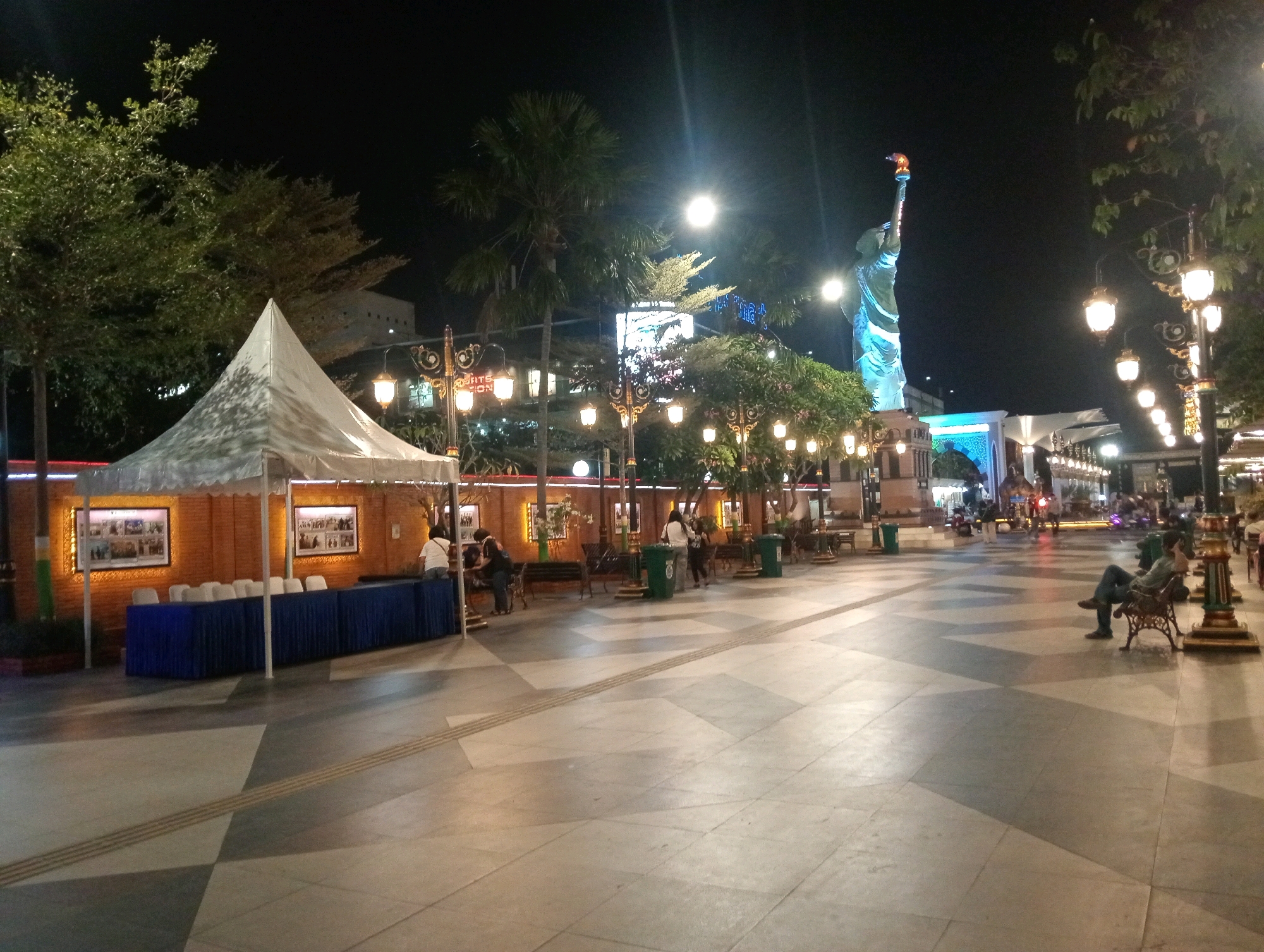 The image shows a night scene at Alun-Alun Pahlawan in Madiun, Indonesia. In the background, a large statue resembling the Statue of Liberty is illuminated and stands tall against the dark sky. The statue is blue and its torch is lit. In the foreground, there is a white tent set up with a long blue table covered with chairs behind it. Two framed pictures hang on the wall behind the tent. Several people are visible in the scene: some are walking, others are sitting on benches, and one person is standing behind the blue table. Decorative streetlights with a vintage design are spaced along the walkway. The ground is paved with large tiles in a geometric pattern. Trees and foliage are also present throughout the scene, adding to the ambiance.