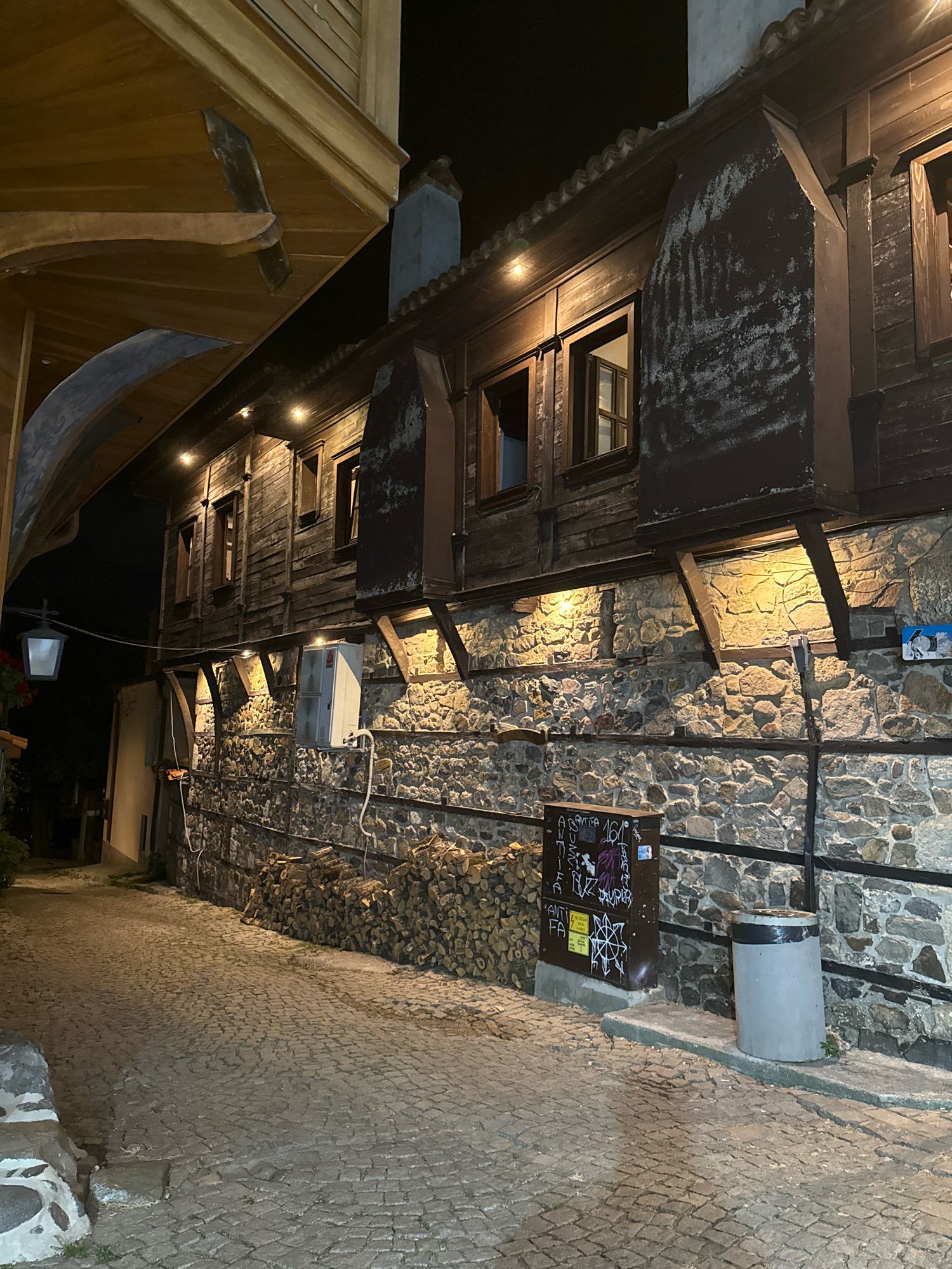 This is a night-time, eye-level, medium shot of a cobblestone street in Sozopol, Bulgaria. The street is flanked by an old, wooden house with stone foundations. The house has several windows, some of which are lit from within, and is illuminated by a few spotlights on the facade. A pile of firewood is stacked against the stone wall. To the right of the firewood, there is a metal utility box covered in graffiti and a cylindrical trash can. A black metal fence runs along the stone wall. The cobblestone street is illuminated by the lights from the house and a vintage-style lantern hanging from an overhead wooden structure on the left side of the frame.