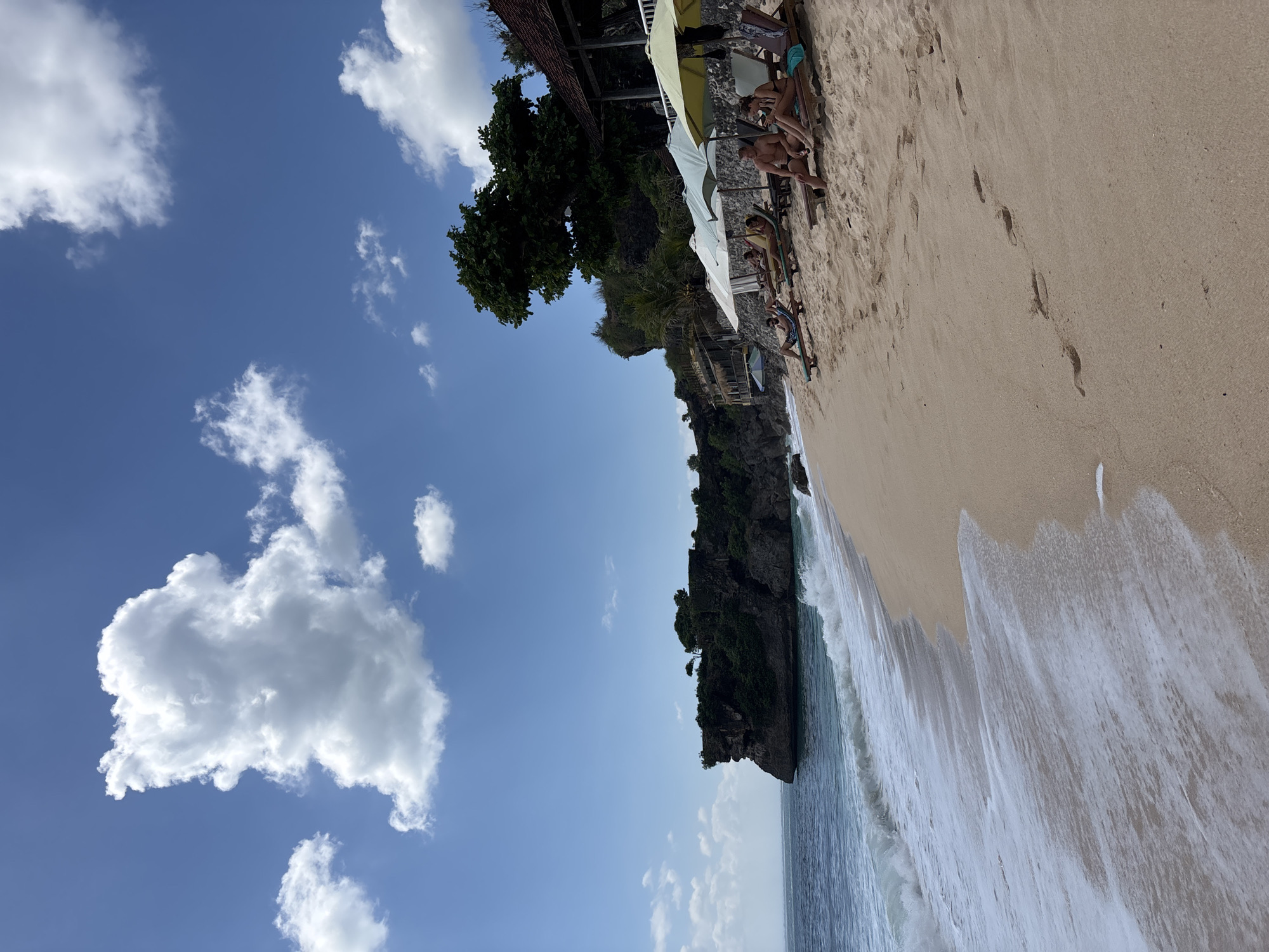 This image captures a wide shot of a sandy beach in Kuta Selatan, Indonesia, under a bright blue sky dotted with scattered white cumulus clouds. To the left, a dark, rugged cliff densely covered in green tropical vegetation extends into the turquoise ocean, where white waves are breaking and receding along the shoreline. The light tan sand of the beach foreground shows multiple sets of human footprints leading from the water's edge towards the mid-ground. Along the right side of the beach, several individuals are visible, relaxing on lounge chairs positioned beneath various colored beach umbrellas (light blue, pale green, yellow-green). Further up the hillside, structures with red-tiled roofs are partially visible amongst the lush greenery. The ocean surface appears calm beyond the breaking waves, meeting the horizon under a clear sky.