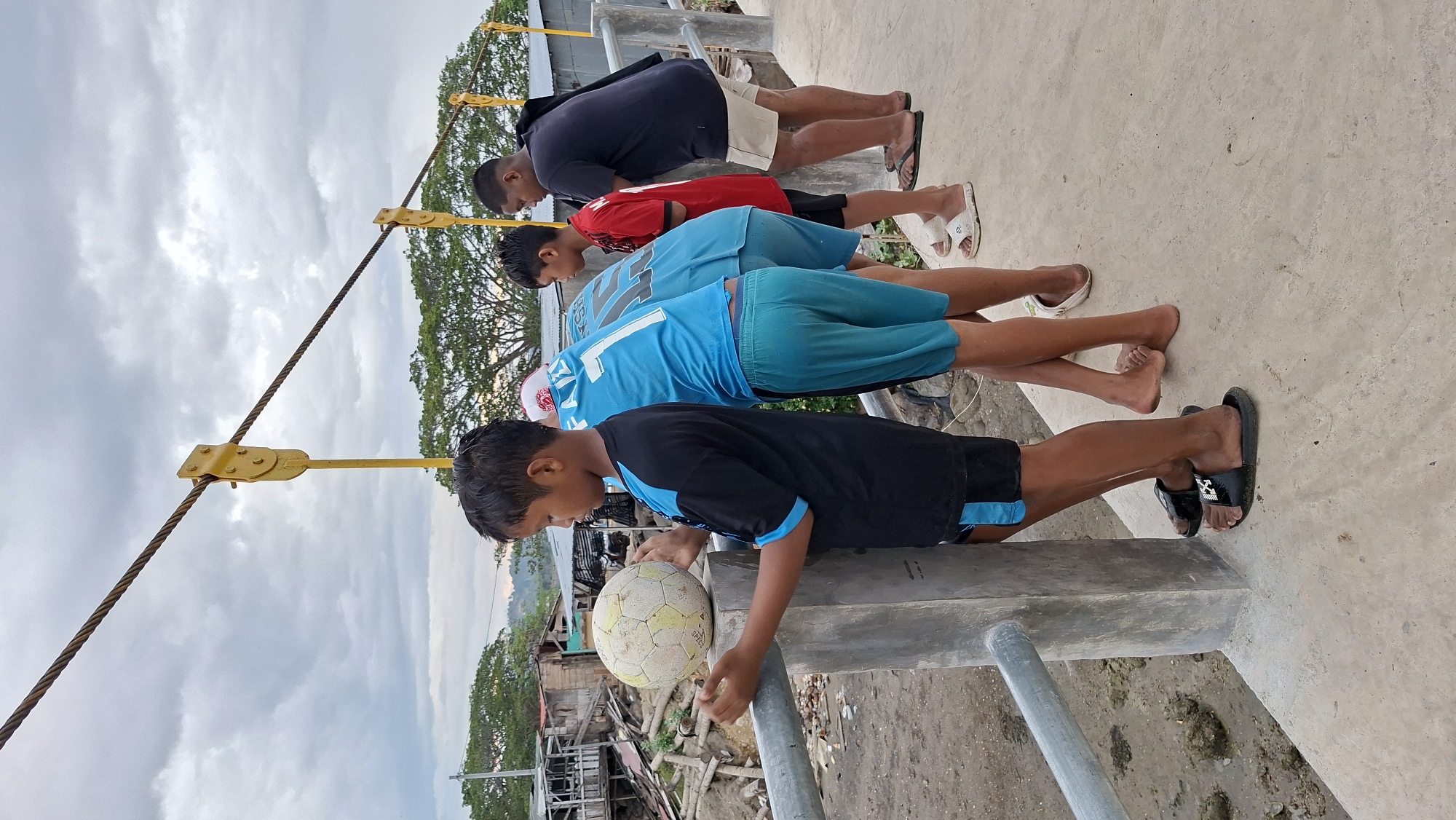 Four young boys stand on a concrete bridge, looking down at the muddy ground below. The boy closest to the viewer, wearing a black and blue shirt, holds a worn soccer ball on a concrete pillar. He is facing left, with his head turned to the side. To his right, three other boys are clustered together. The second boy from the left wears a blue jersey with the number "1" prominently displayed. The third boy from the left is in a red shirt, and the fourth boy, on the far right, is wearing a dark blue t-shirt and light-colored shorts. They are all looking downwards, with their stances suggesting they are observing something.
The environment appears somewhat disheveled. The ground below the bridge is muddy and appears to have debris, including what looks like discarded wood and construction materials, suggesting a potentially degraded or informal settlement area. In the background, there are structures that resemble modest housing or temporary buildings, some with corrugated metal roofs. Trees are visible further in the background, and the sky is overcast with dramatic clouds. The bridge itself has a metal railing, and large, thick cables run overhead, attached to yellow vertical supports. The scene suggests a moment of casual observation or perhaps a pause in play on a cloudy day in an urban or semi-urban setting in Lhokseumawe, Indonesia.