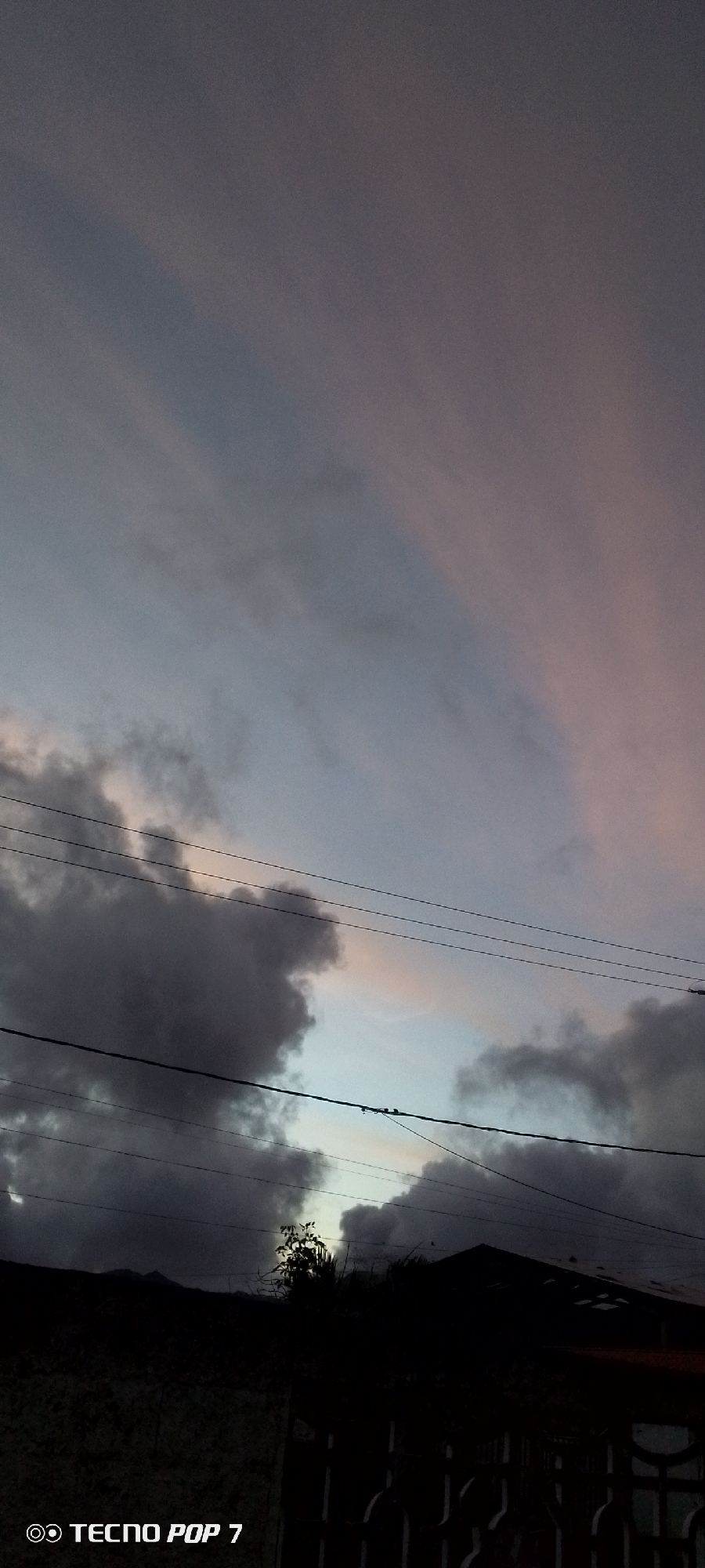 This image depicts a cloudy sky at either dawn or dusk, set in Altagracia, Venezuela. The sky transitions from soft pink and purplish-grey clouds in the upper portion to lighter blue-grey tones near the horizon. Dense, dark grey clouds are prominent in the mid-ground, creating a dramatic, overcast atmosphere.
Several utility lines, likely power or communication cables, traverse the frame horizontally, adding a man-made element to the natural skyscape. The foreground is almost entirely in silhouette, featuring the dark outlines of structures, possibly houses or buildings, on the right with a discernible roofline and a patterned fence or gate. A cluster of trees and foliage, also in silhouette, stands out against the slightly brighter part of the sky in the lower-middle, with minimal detail visible due to the low light. Faint, distant silhouettes of mountains or hills are visible along the lower-left horizon, suggesting a varied landscape.
No people or explicit activities are visible, conveying a sense of quietude and stillness. The soft, fading light and deep shadows contribute to an atmospheric and tranquil mood. In the bottom-left corner, the text "TECNO POP 7" with a camera icon is visible, indicating the device used to capture the image.