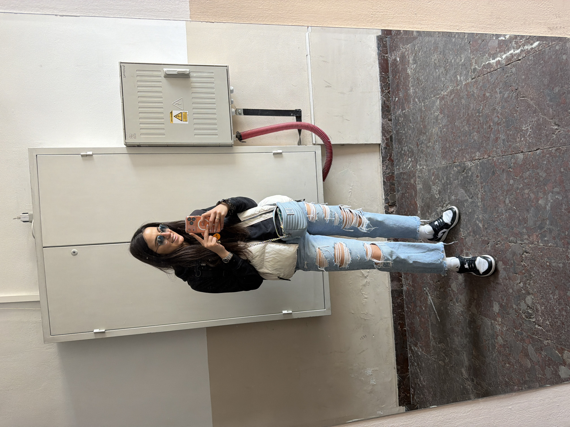 A young woman is reflected in a large metal door, likely of an electrical box, indoors. She is taking a selfie with a smartphone. She wears blue sunglasses, a black jacket, and ripped blue jeans. A white handbag hangs across her body. She stands on a floor covered in marble tiles. The scene is illuminated by artificial light. Above the electrical box is another box with a warning symbol. A flexible red pipe connects to the lower box. The location is likely within a public building in Sofia, Bulgaria.