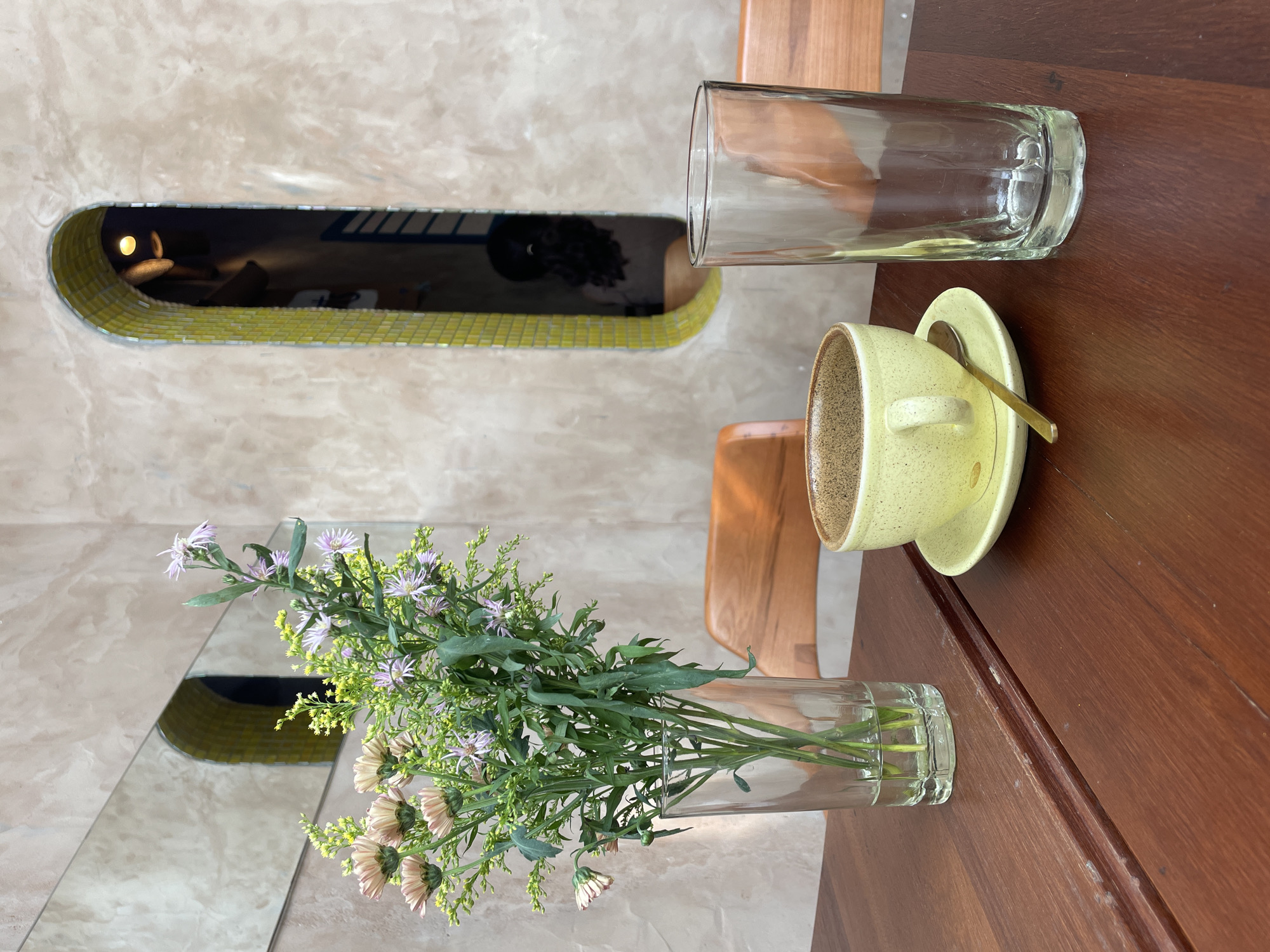 A high-angle, medium shot captures a wooden table at what appears to be a cafe in Mũi Né, Vietnam. The table surface is a rich, dark wood with visible grain and some wear. To the left, a tall, clear glass vase holds a bouquet of light pink and yellow flowers with green stems and leaves. In the center, a pale yellow ceramic coffee cup sits on a matching saucer, accompanied by a gold spoon. To the right of the cup, an empty, clear glass tumbler stands tall. The background features a textured wall with an arched niche, intricately tiled in yellow mosaic. A mirror above the niche reflects the interior, showing a dimly lit space with a curved, yellow-tiled archway and a glimpse of a person with dark hair. Wooden chair backs are visible on either side of the table. The overall ambiance suggests a calm and slightly artistic setting.