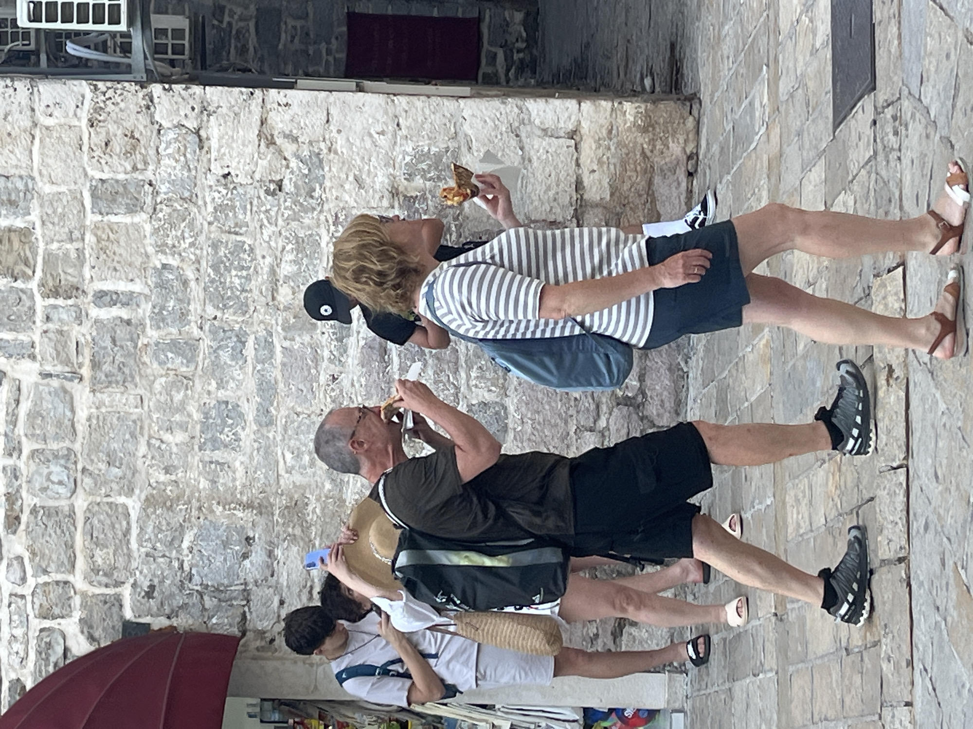 In Budva, Montenegro, a group of people are walking on a cobblestone street in front of a stone building. A man in a dark t-shirt and black shorts is eating a slice of pizza while walking. Beside him, a woman with blonde hair wearing a striped t-shirt and dark shorts is also holding and eating a slice of pizza. Behind them, a young person with dark hair wearing a white t-shirt and shorts is holding a phone and a straw hat. Another person in a black cap is walking behind them. The environment is an outdoor urban setting with a stone wall and buildings in the background.