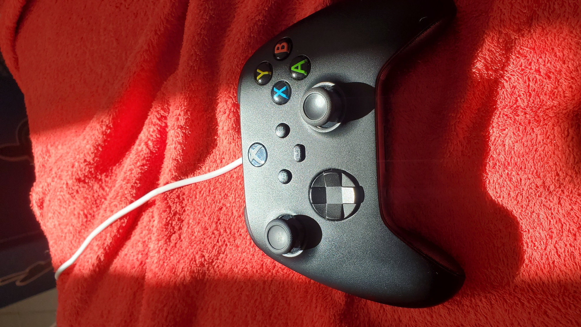 A black Xbox Series X/S controller, featuring two analog sticks, a checkered D-pad, ABXY buttons (green A, red B, blue X, yellow Y), menu, view, and a central illuminated Xbox logo button, rests on a vibrant red, textured blanket or towel. A white USB-C cable is connected to the top of the controller, extending upwards and out of the frame. Strong natural light, consistent with direct sunlight, illuminates the scene from the upper left, creating a prominent diagonal shadow across the red material and partially obscuring the right side of the controller. No individuals are present, and no actions or interactions are depicted. The scene is observed in an indoor setting within Curitiba, Brazil.