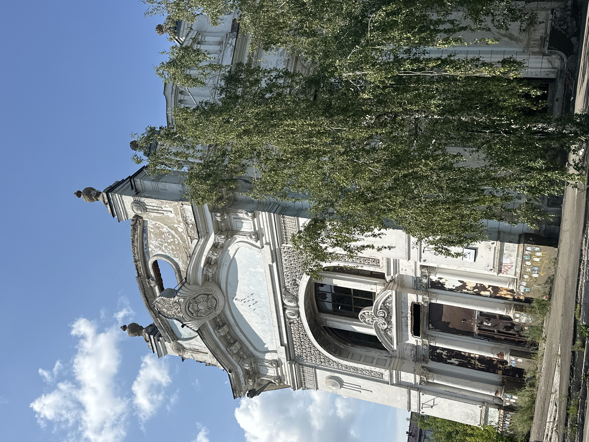 The image captures the exterior of a dilapidated building in Biysk, Russia, on a clear day with scattered clouds. The building, likely an old public structure, exhibits ornate, classical architectural details that are now weathered and damaged. The facade is predominantly white, with peeling paint and visible signs of decay, including crumbling plaster and rust stains around the doors and windows.
A prominent feature is a large, arched entryway with heavy, corroded metal doors. Above this, a large window with broken panes is visible, surrounded by intricate stucco work. Decorative elements like columns, moldings, and finials adorn the facade and the roofline, hinting at its former grandeur. A large oval opening is present on the upper part of the building, possibly a former window or decorative element, now exposed to the elements.
A mature birch tree with abundant green foliage stands to the right of the building, partially obscuring the facade and reaching towards the roof. The ground in front of the building is paved, with overgrown weeds and grass sprouting from cracks and along the edges, suggesting neglect. Faded posters or graffiti are affixed to the lower sections of the building walls. In the background, glimpses of other urban structures and trees can be seen, indicating an urban or semi-urban setting. The overall impression is one of abandonment and the slow decay of a once-significant building.