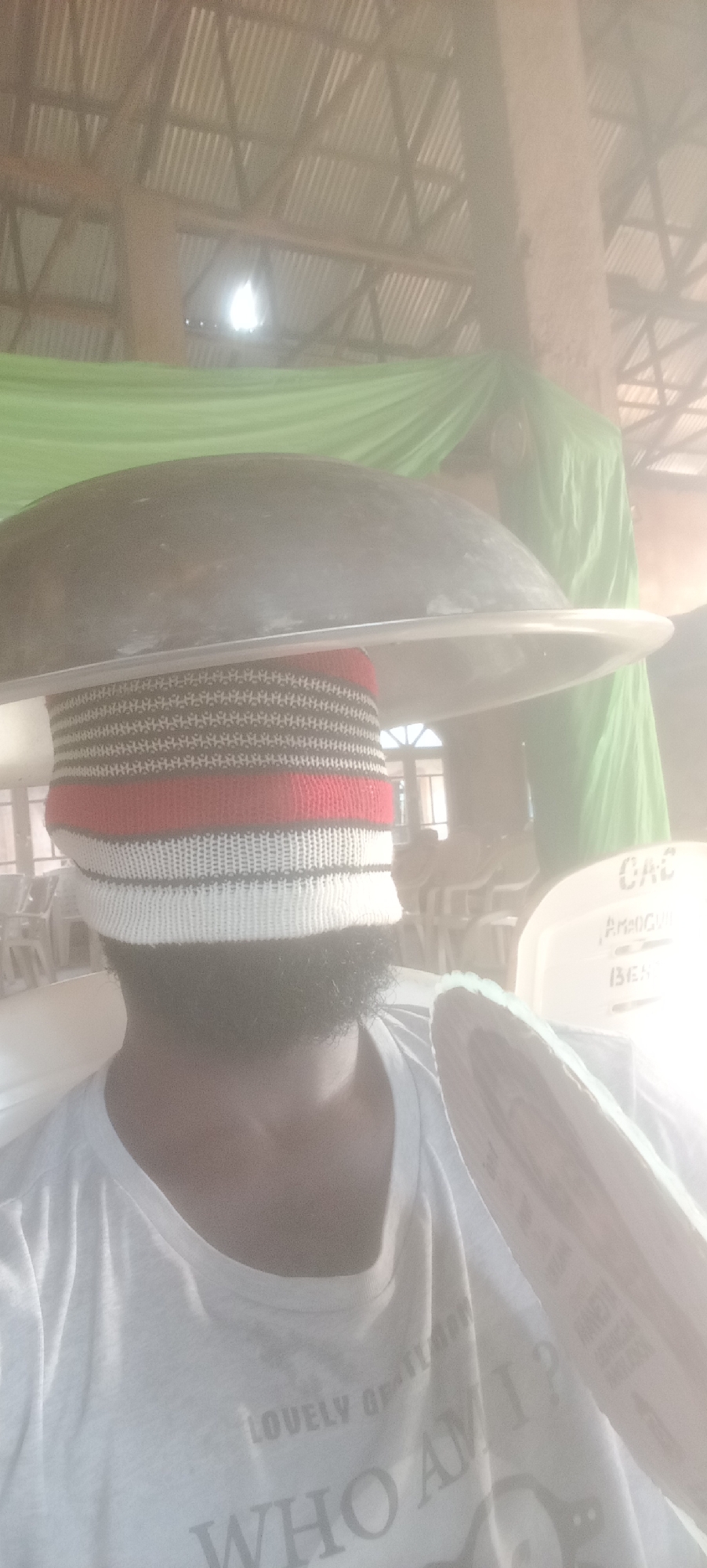 A man with a beard is wearing a knit cap covering his eyes, with a large, dark pan resting on top of his head. He is wearing a white t-shirt with the words "LOVELY GENTLEMAN" and "WHO AM I?" printed on it. The background shows what appears to be an interior space, possibly a church or event hall, with rows of white chairs, green draped fabric, and a corrugated metal roof. The environment is dusty or hazy. A chair in the background reads, "CAC AMOGU BENDE". The scene is taking place in Bende, Nigeria.