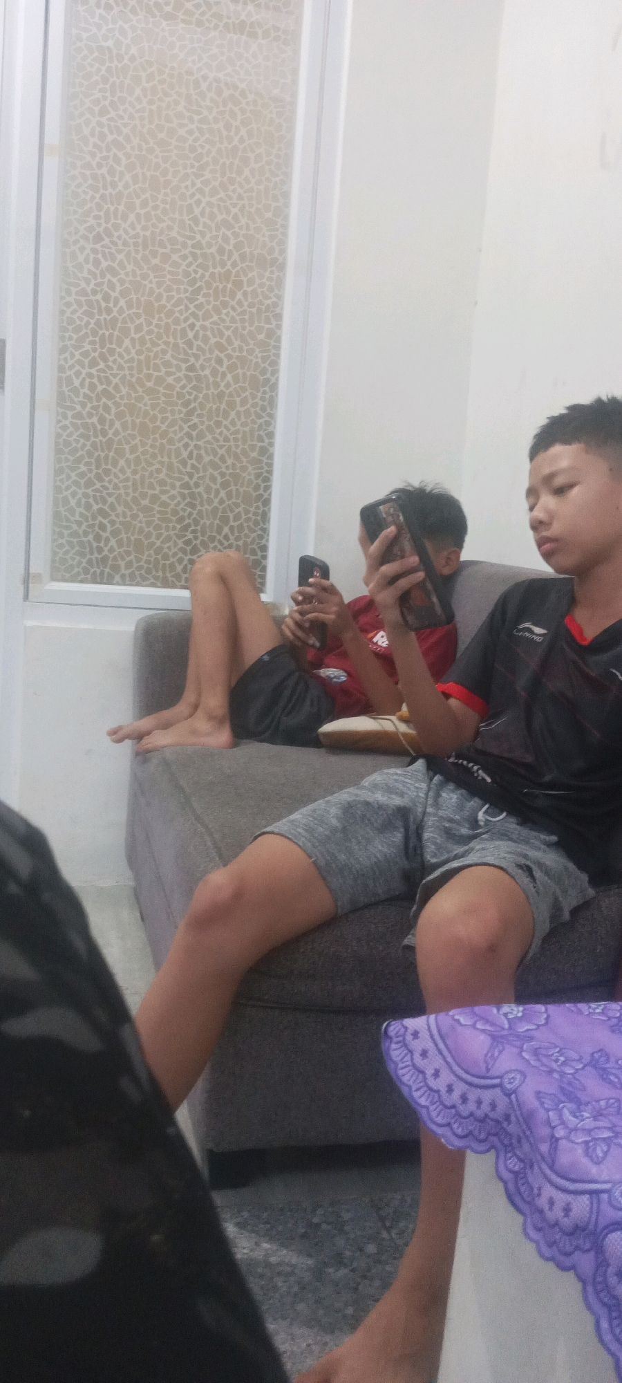 The image depicts two young males sitting on a grey sofa. The male on the left is wearing black shorts and a red t-shirt, and is holding a mobile phone. The male on the right is wearing grey shorts and a black t-shirt with red accents, and is also holding a mobile phone. They are positioned in a room with a white wall and a window with a textured glass pattern. A purple patterned cloth is visible on the lower right side of the image. The scene appears to be indoors, possibly in a residential setting within Babelan, Indonesia.
